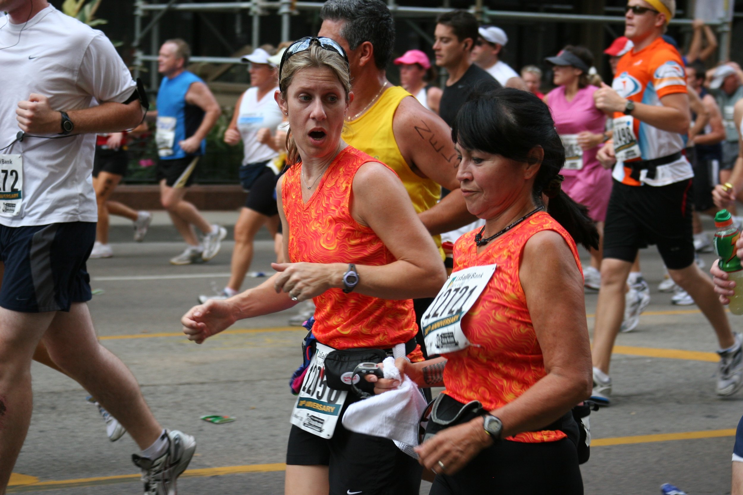 Women in bright orange athletic shirts running at a marathon race, with multiple runners and spectators in the background.