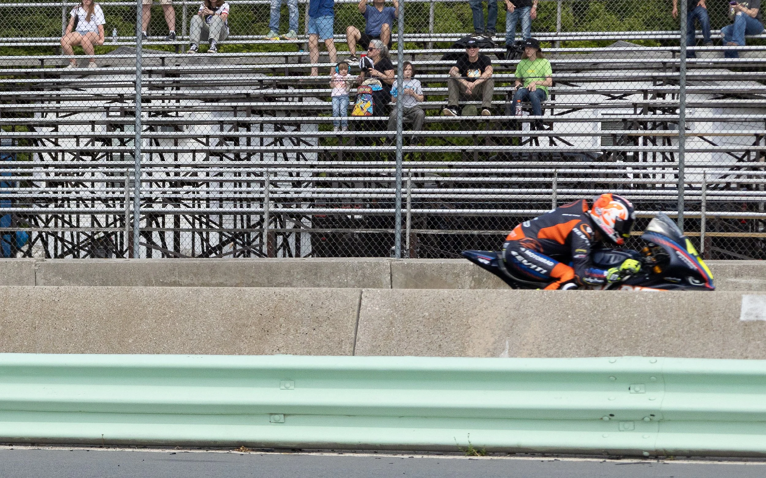 A motorcycle racer in an orange and black suit leaning into a turn on a racetrack, with a chain-link fence and a seated spectators in the background, including children and adults.