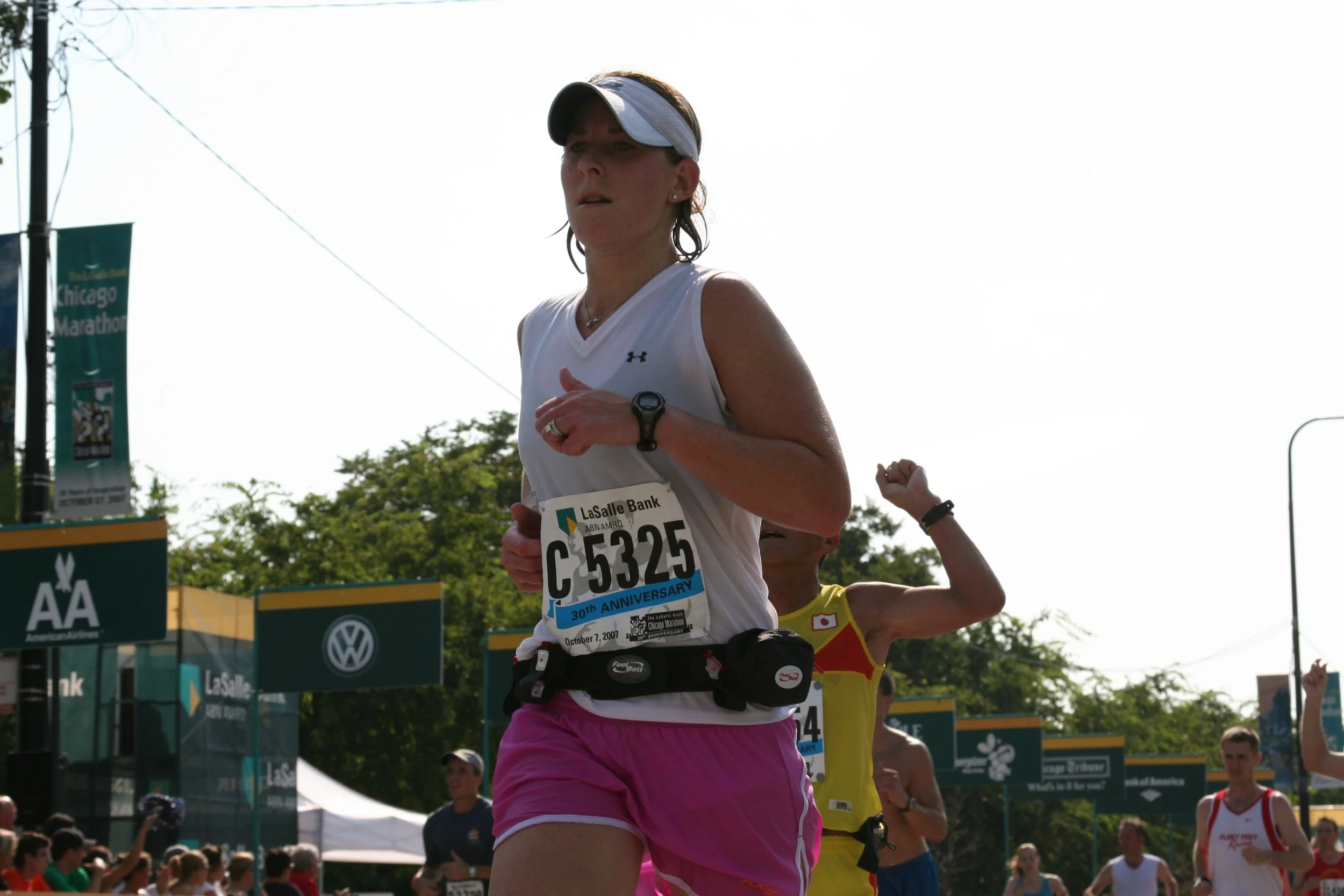 A woman running in a marathon, wearing a white tank top, pink shorts, a white visor, and a race bib, with other runners and marathon banners visible in the background.