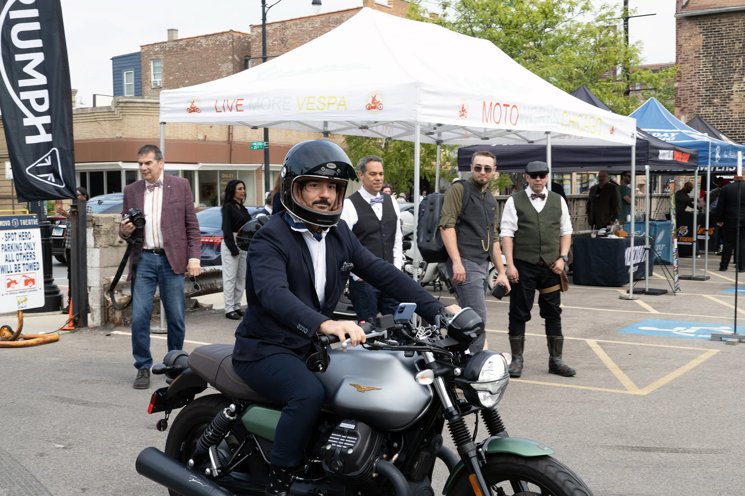 A man in a suit and helmet riding a gray motorcycle at an outdoor event with tents and people in the background.