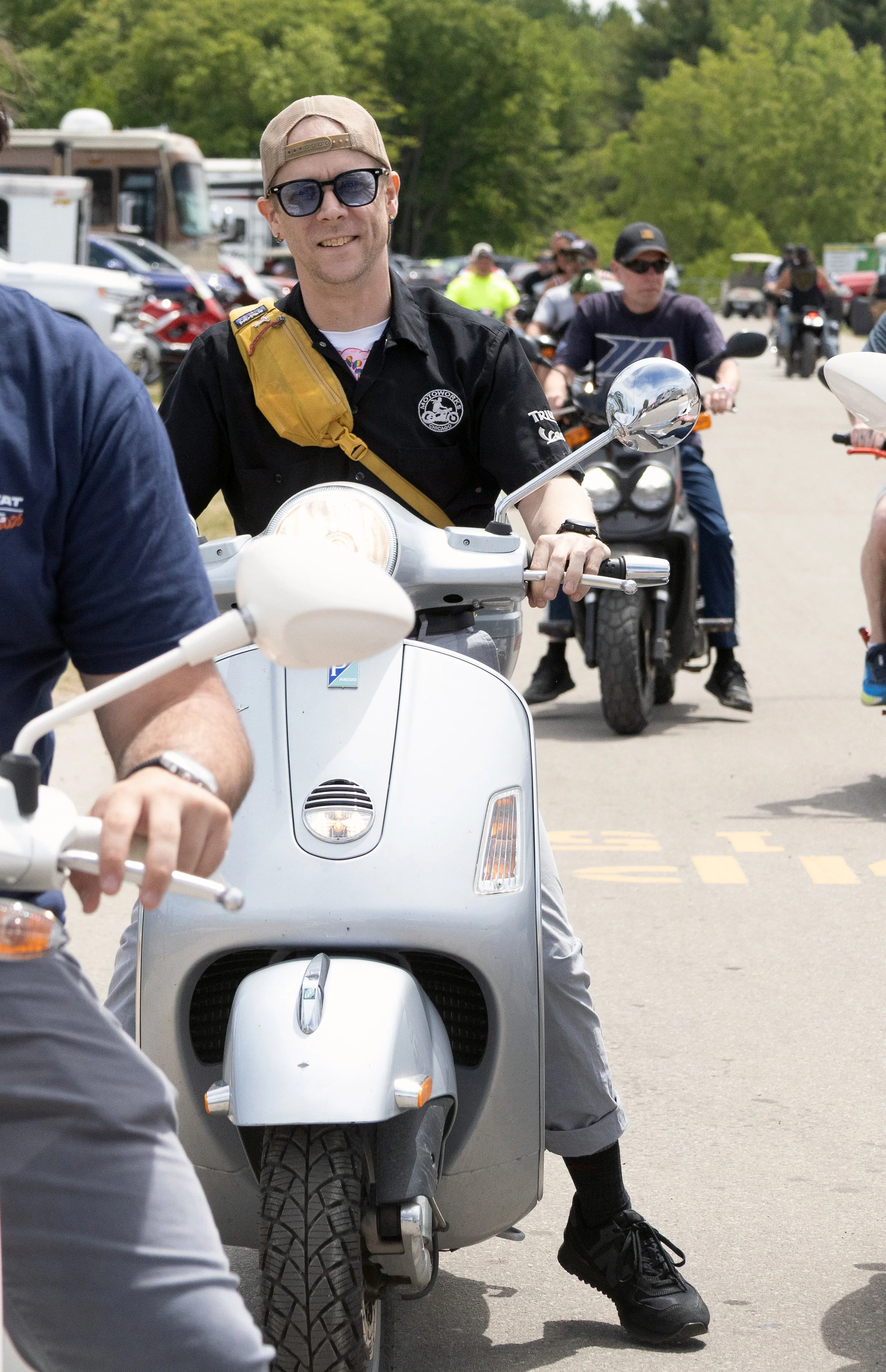 A group of people, including a man in a black shirt with sunglasses and a tan cap, riding scooters in a parking lot surrounded by cars and motorcycles, with green trees in the background.