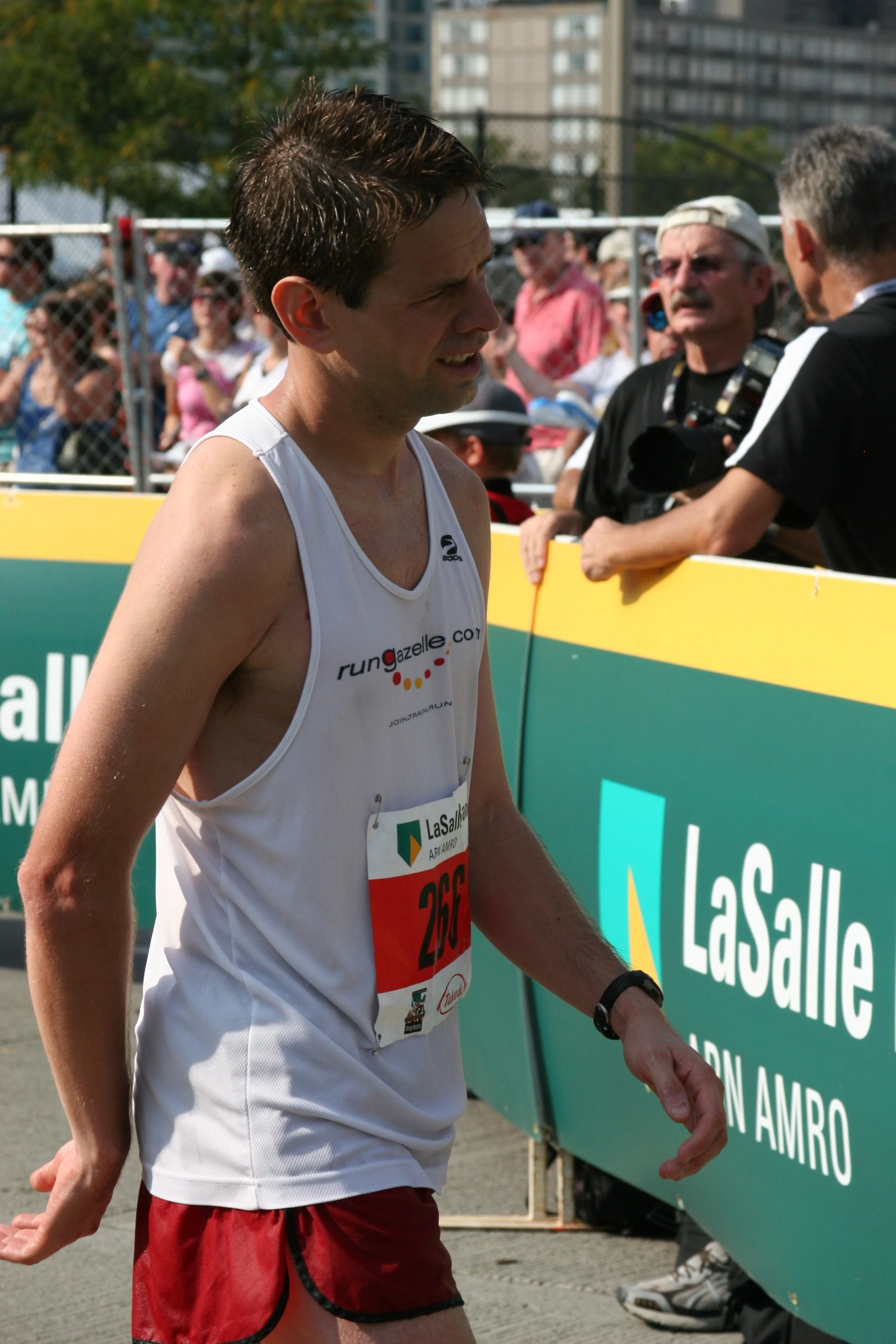 Young male marathon runner wearing a white tank top and red shorts, walking past spectators and a green advertising banner at the race event.