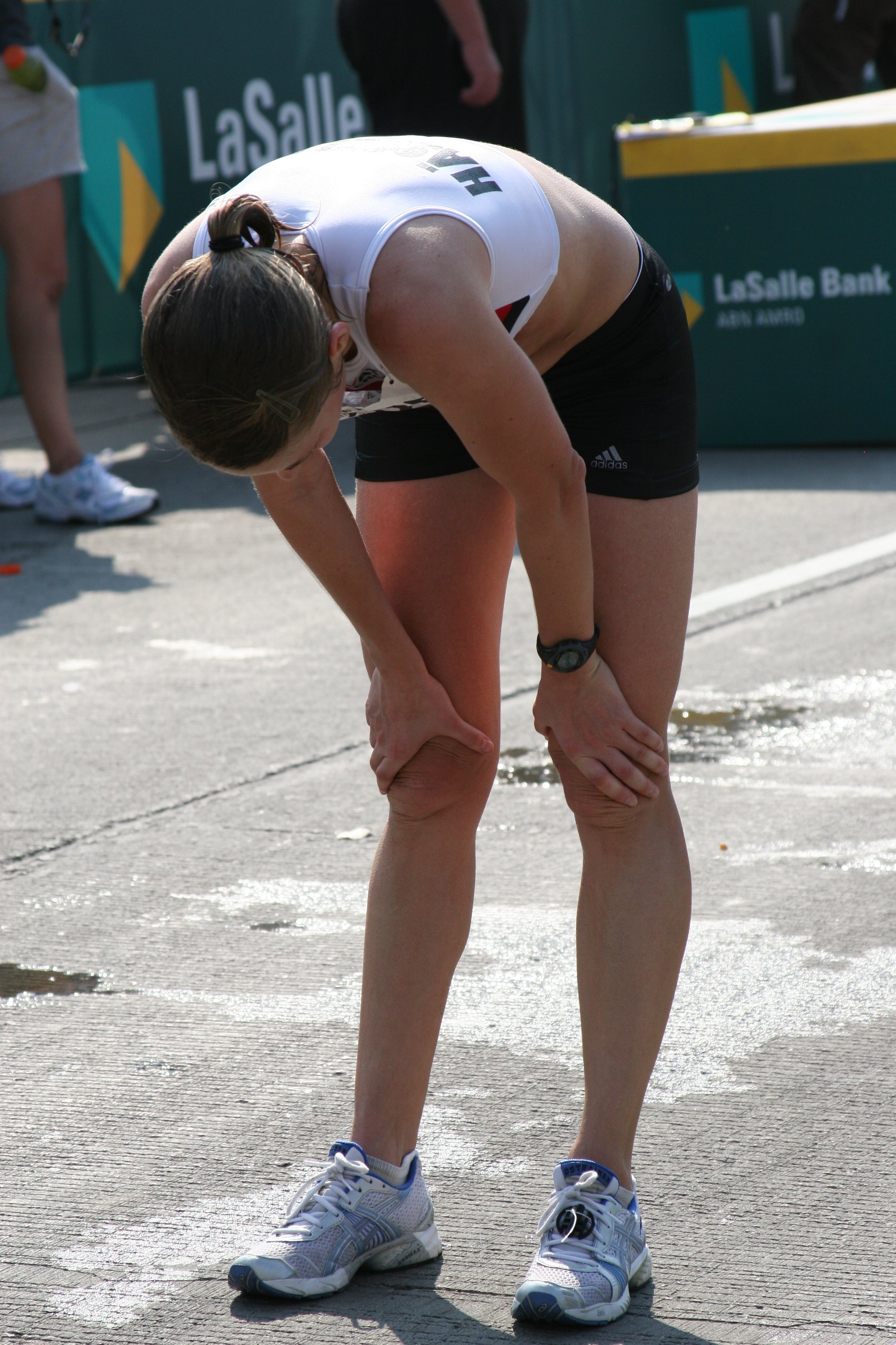 A female athlete in sportswear bent over on a track, resting or catching her breath, with a LaSalle Bank sponsorship banner in the background.