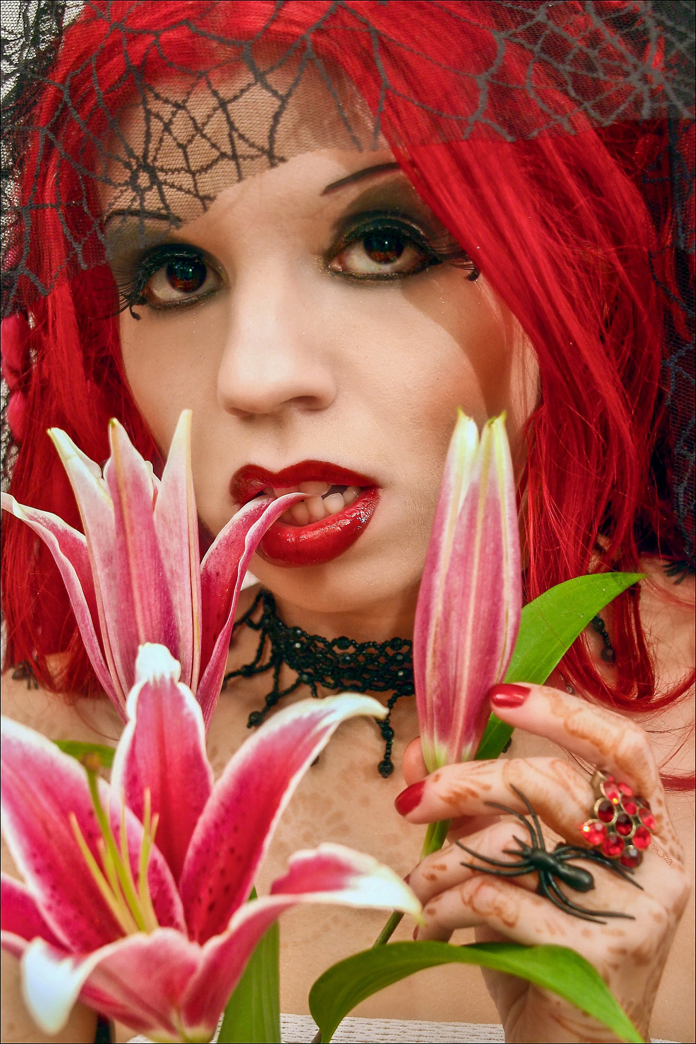 A woman with red hair covered with black lace, wearing dark makeup and red lipstick, holding pink and white lilies, and has a black spider ring on her finger.