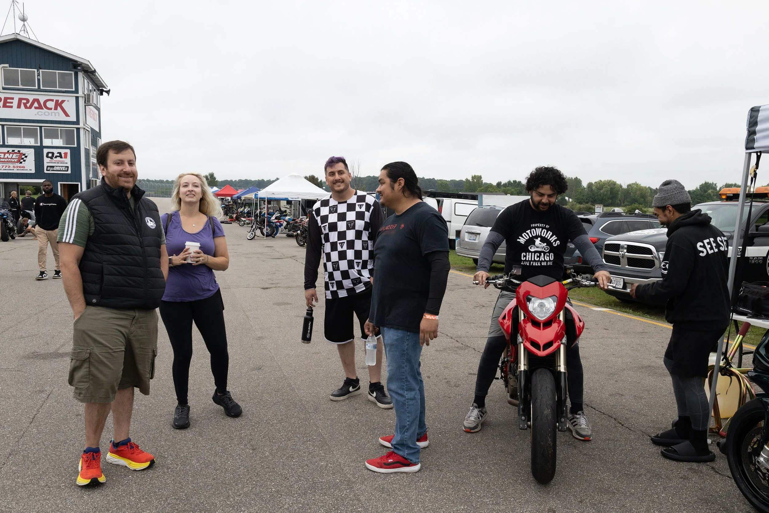 A group of five people standing outdoors at a motorsports event, with tents, motorcycles, and cars in the background. The overcast sky suggests cloudy weather.