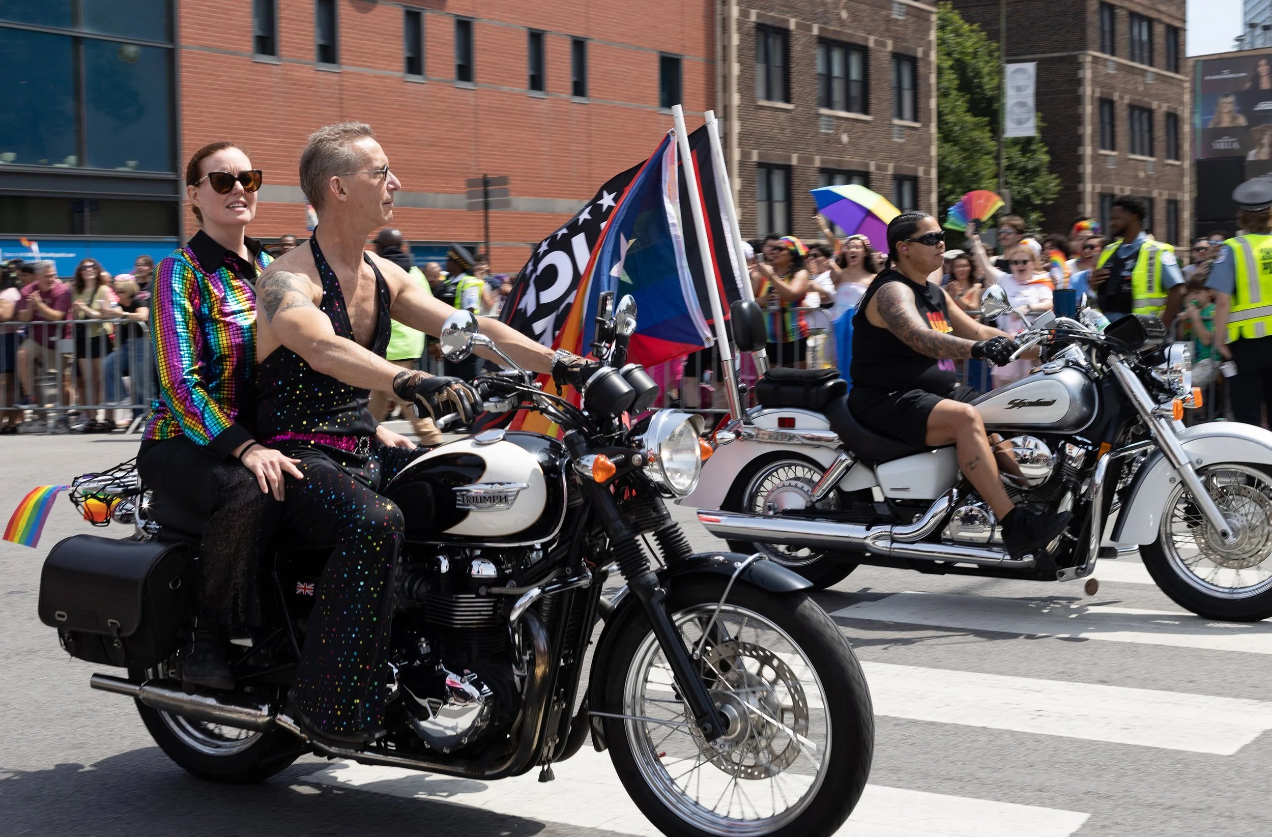 People participating in a pride parade, riding motorcycles decorated with rainbow flags, in a city street lined with spectators and buildings.