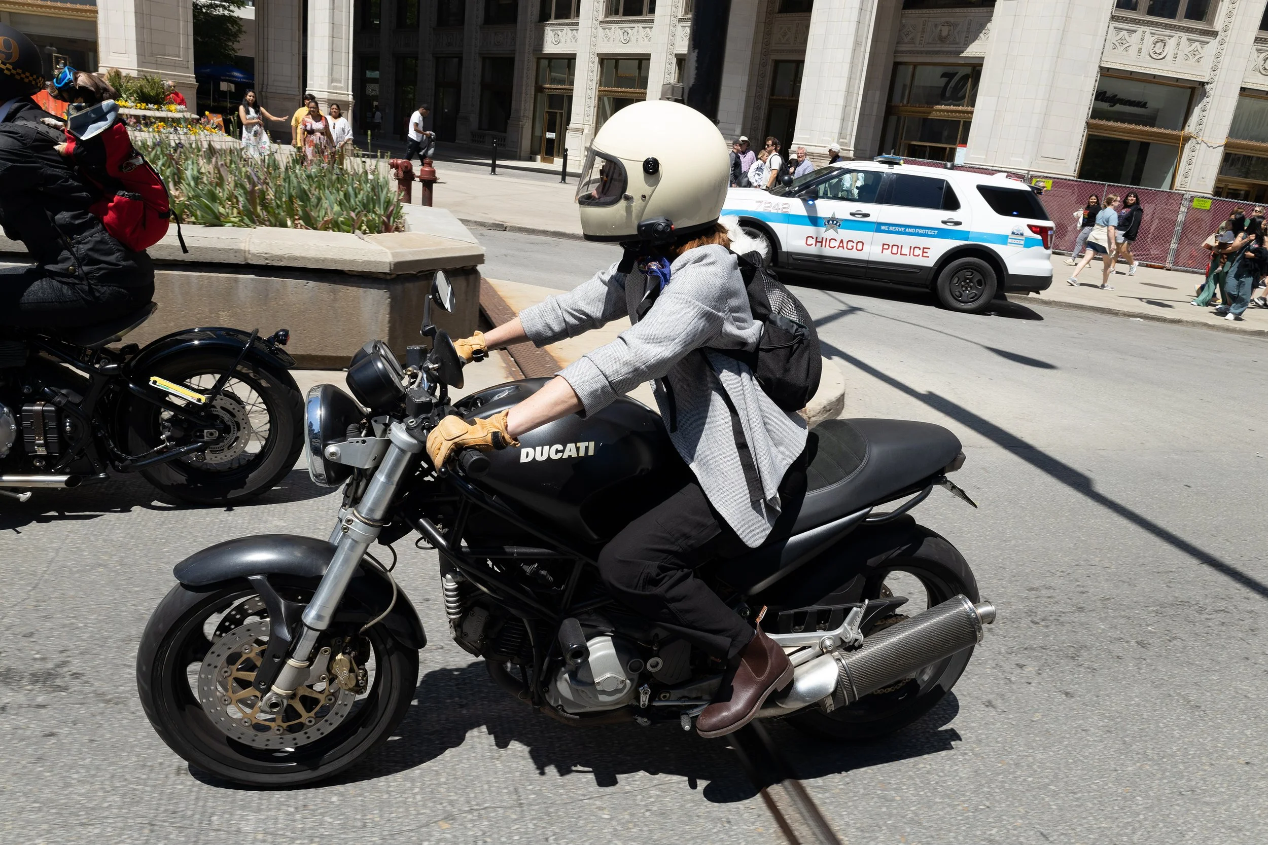 A person riding a black Ducati motorcycle on a city street, wearing a helmet, gray jacket, and gloves. In the background, there are pedestrians walking on the sidewalk, a Chicago police car parked near a building, and a flower bed with green plants.