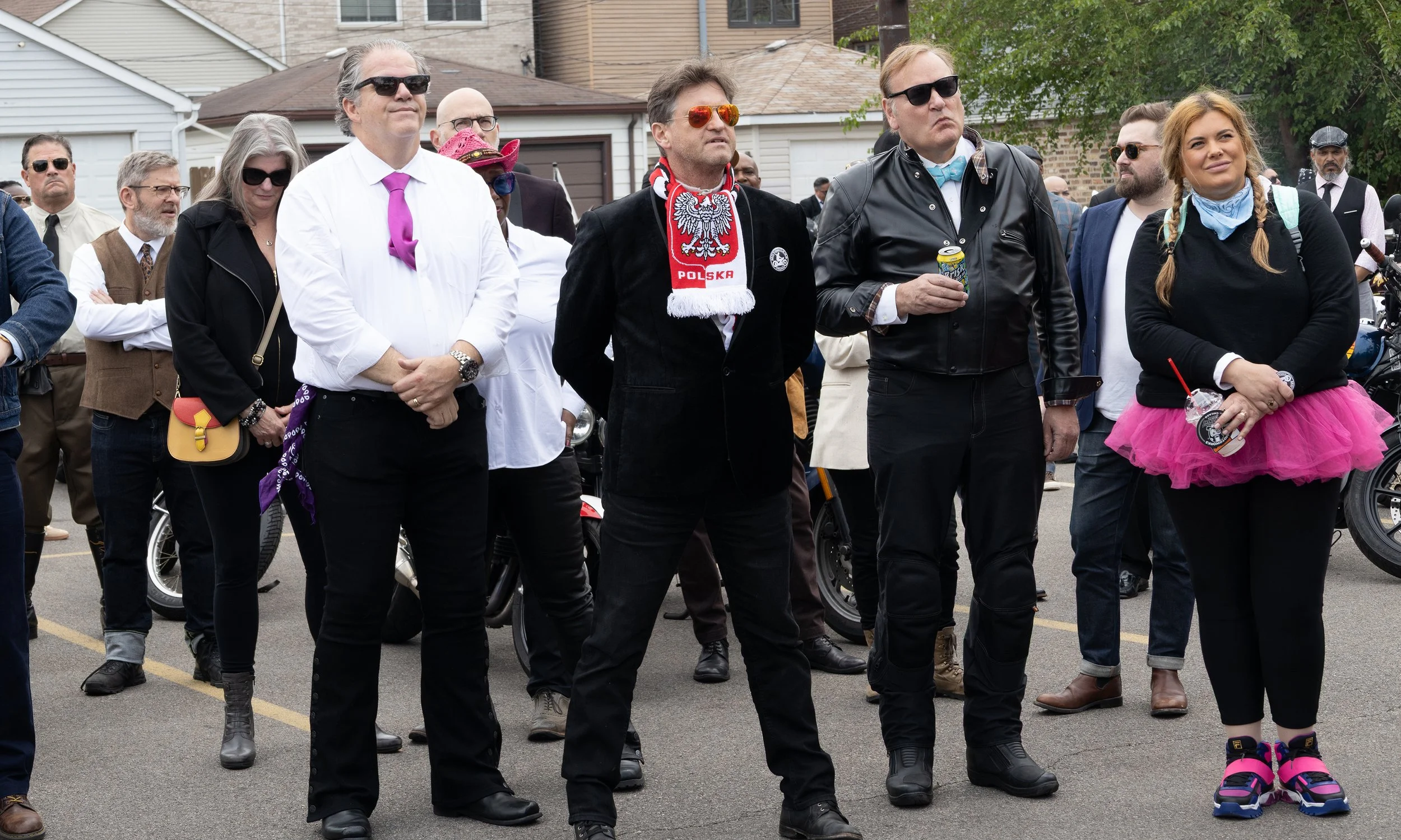 A group of people standing outdoors, dressed in casual and punk-style clothing, some wearing sunglasses, with a background of houses and trees.