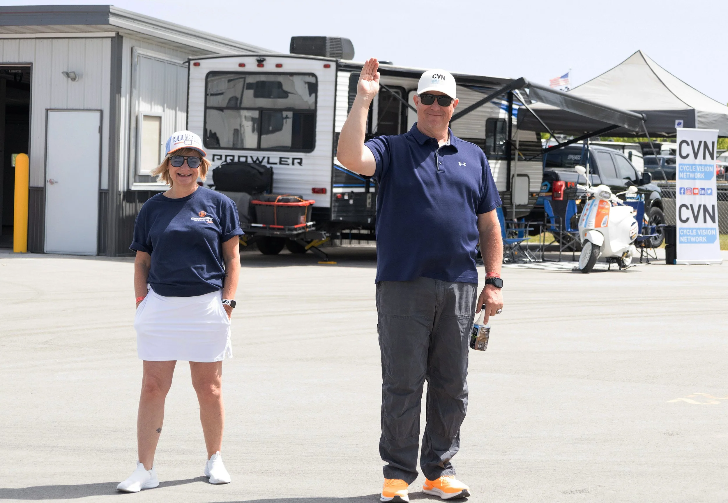 Two smiling individuals waving at the camera in front of a RV and tents, with a sign for Cycle Vision Network and a scooter in the background.