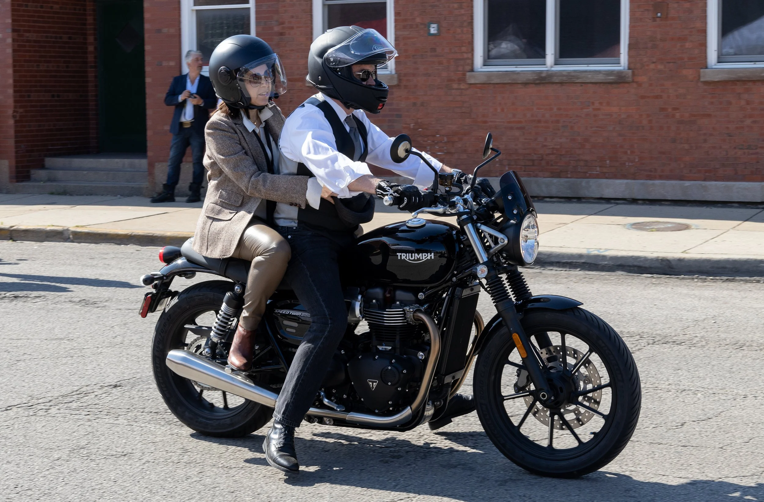 Two people riding a black Triumph motorcycle, both wearing helmets, with a brick building in the background.