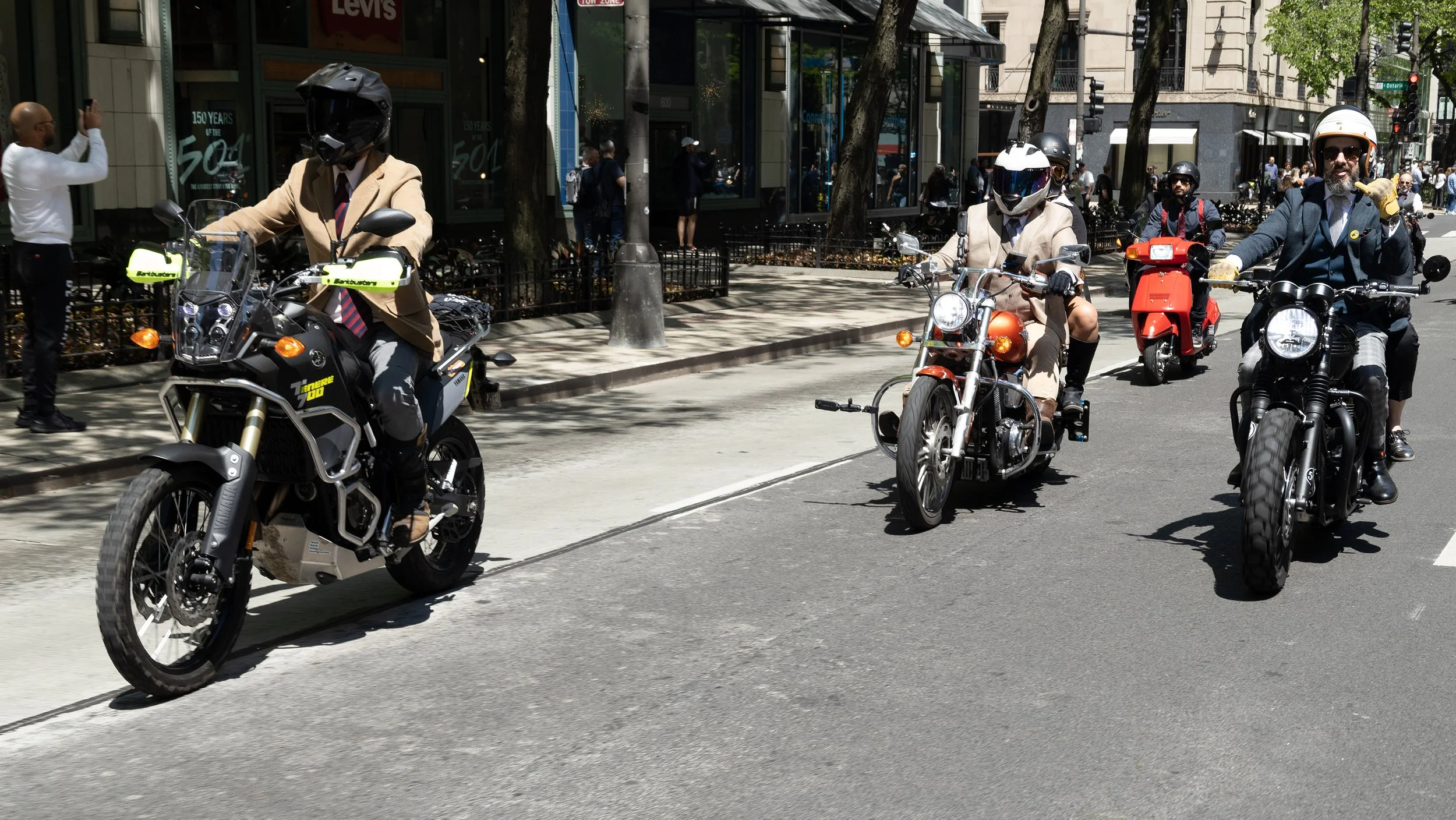 People riding motorcycles and scooters on a city street during daytime, with onlookers on the sidewalk and storefronts in the background.