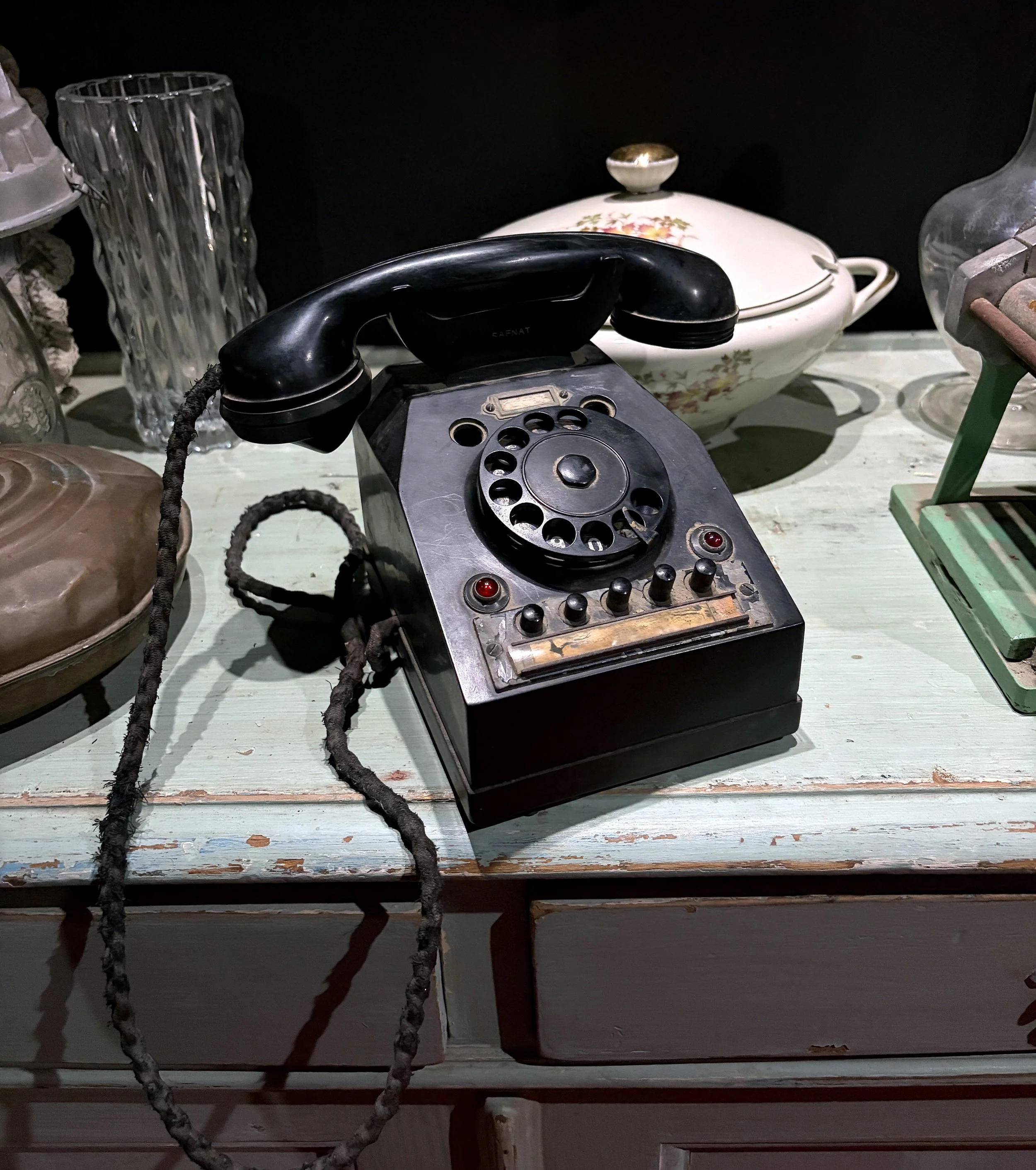 An antique black rotary telephone on a wooden surface with a clear glass vase, a floral-patterned dish with a lid, and other vintage items around.