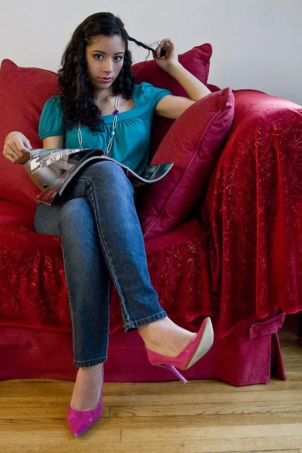 A woman sitting on a red sofa, reading a magazine, wearing a blue top, jeans, and pink high heels, with one hand on her hair.