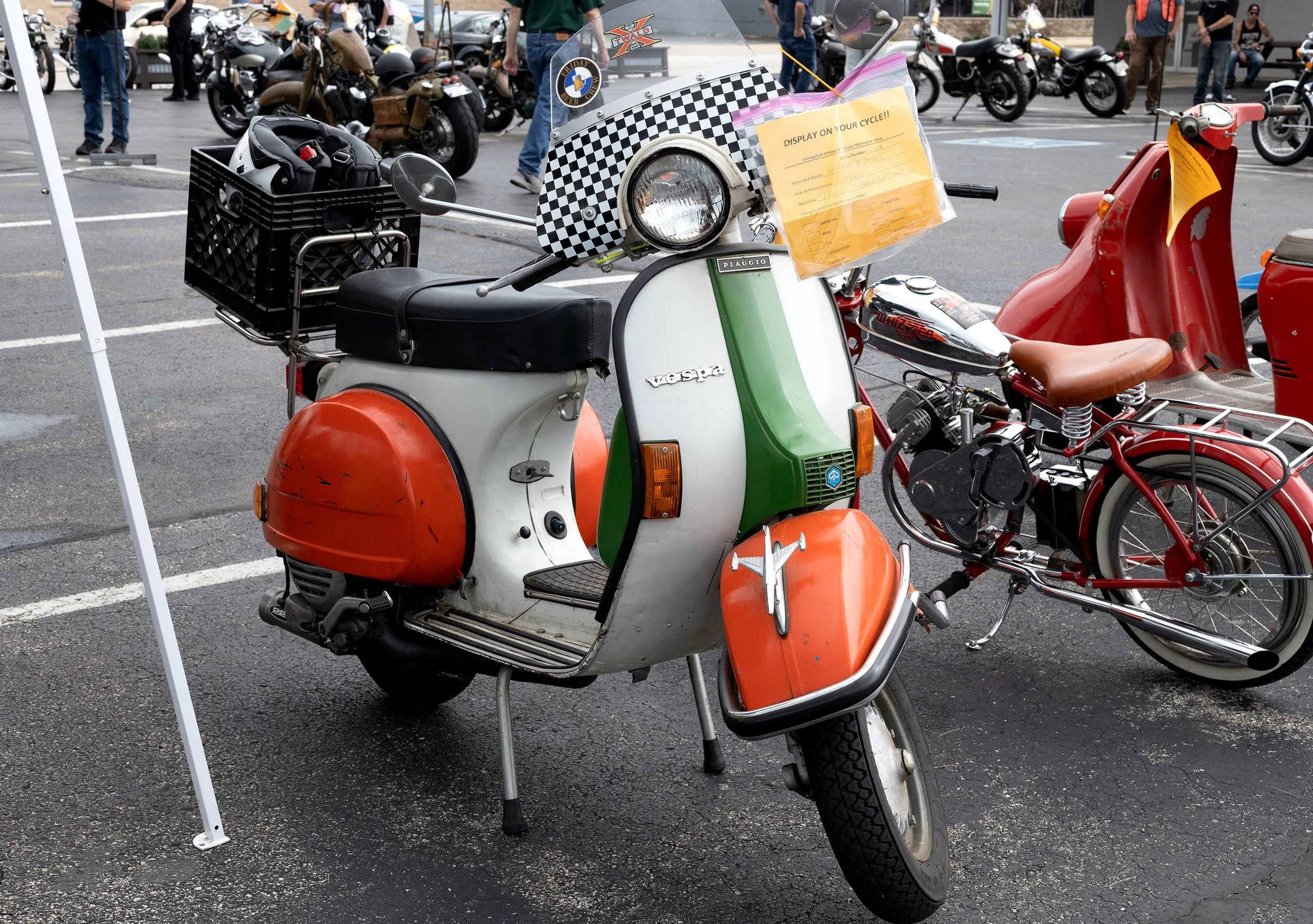 A vintage Vespa scooter with a checkered pattern front fender, a yellow display sheet, a black basket with a helmet, and other motorcycles visible in the background at an outdoor motorcycle show.