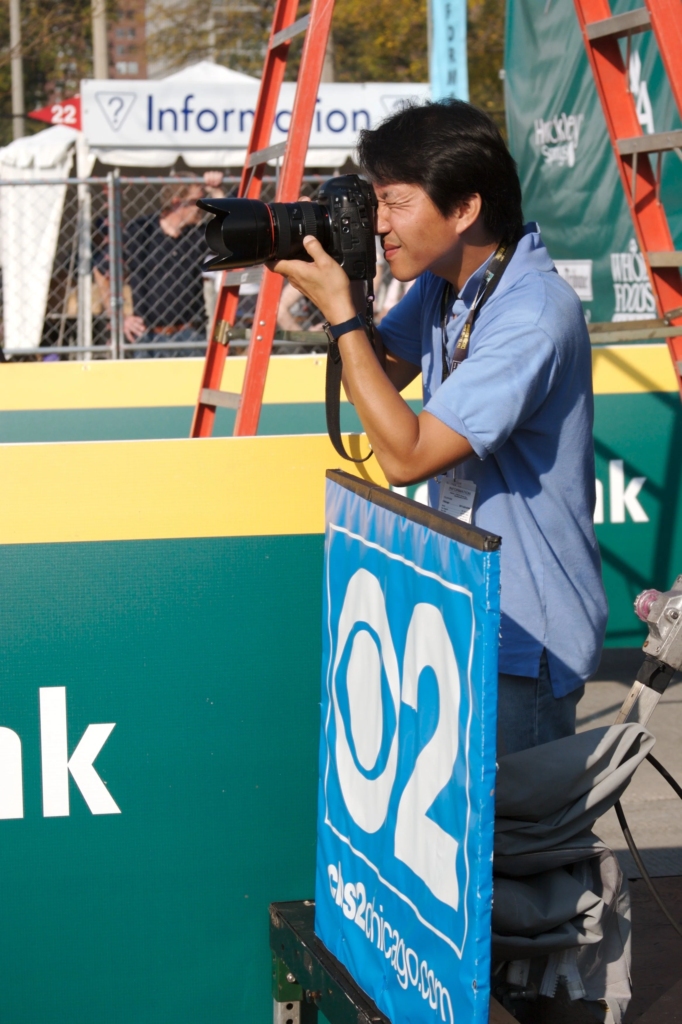 Photographer with a professional camera takes pictures at an outdoor event. Background includes a white tent with a sign and a person in front of a fence.