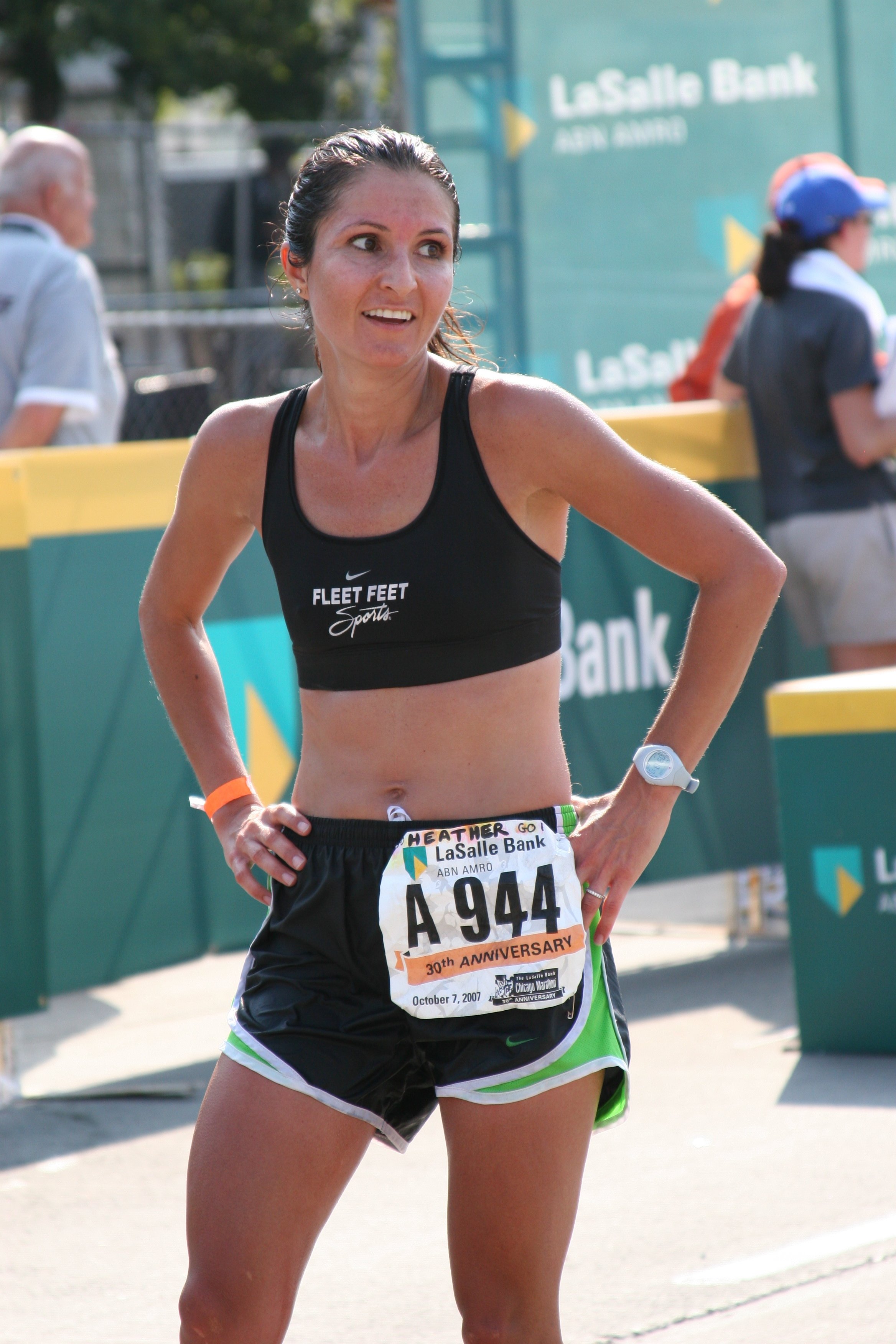 A female marathon runner wearing a black sports bra and shorts, with a race bib showing the number 944, stands with her hands on her hips during a race. She appears sweaty and focused, with other runners and a green banner for LaSalle Bank in the bac