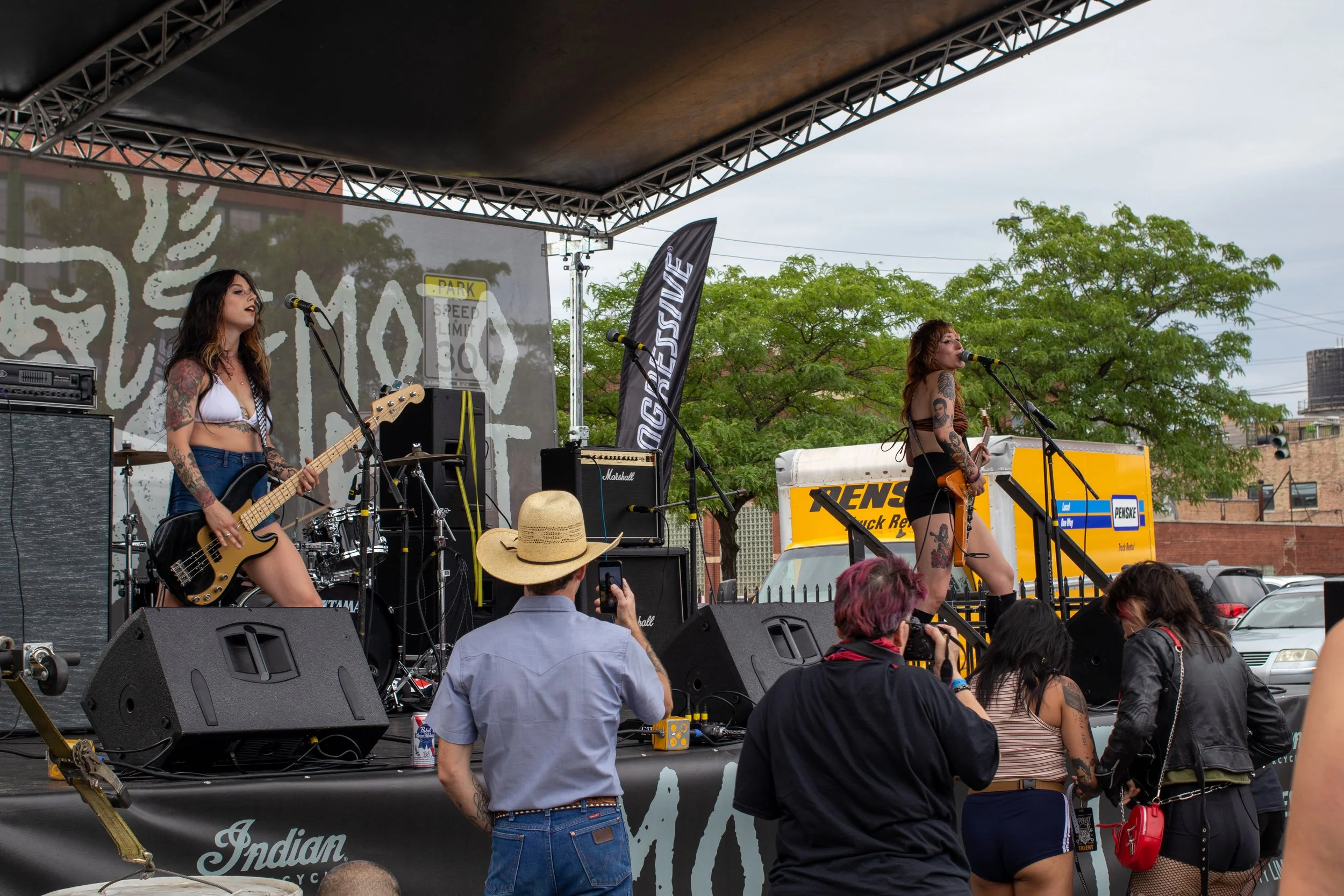 Two female musicians performing on an outdoor stage with musical equipment and an audience watching. One is playing a bass guitar and the other is singing into a microphone, with green trees and buildings in the background.