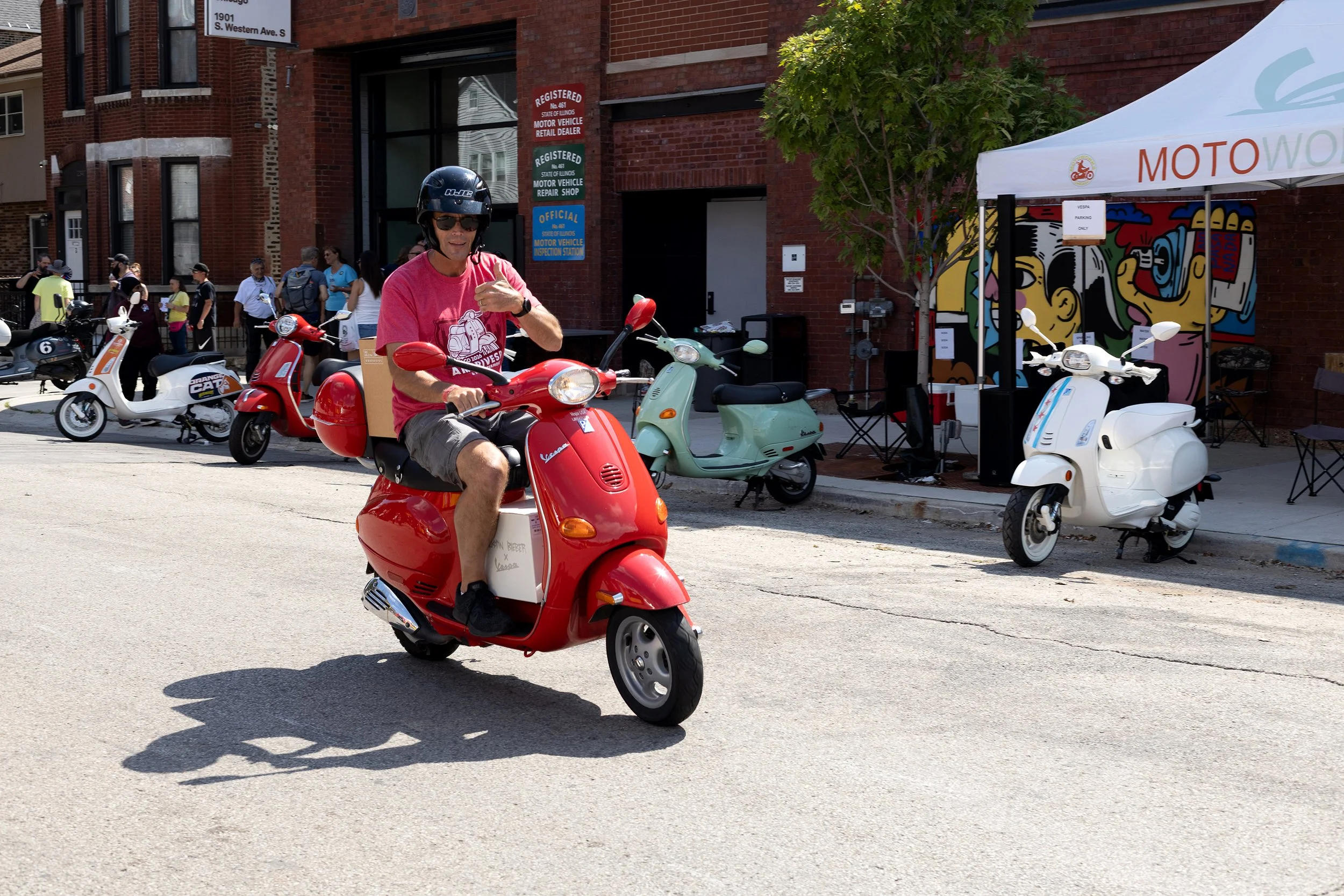 A man wearing a helmet and sunglasses riding a red Vespa scooter on a city street with multiple scooters parked and a group of people in the background.