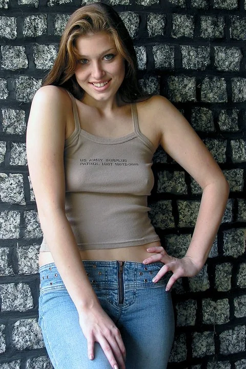 A young woman with long brown hair wearing a beige tank top and blue jeans, standing against a textured gray brick wall, smiling at the camera.
