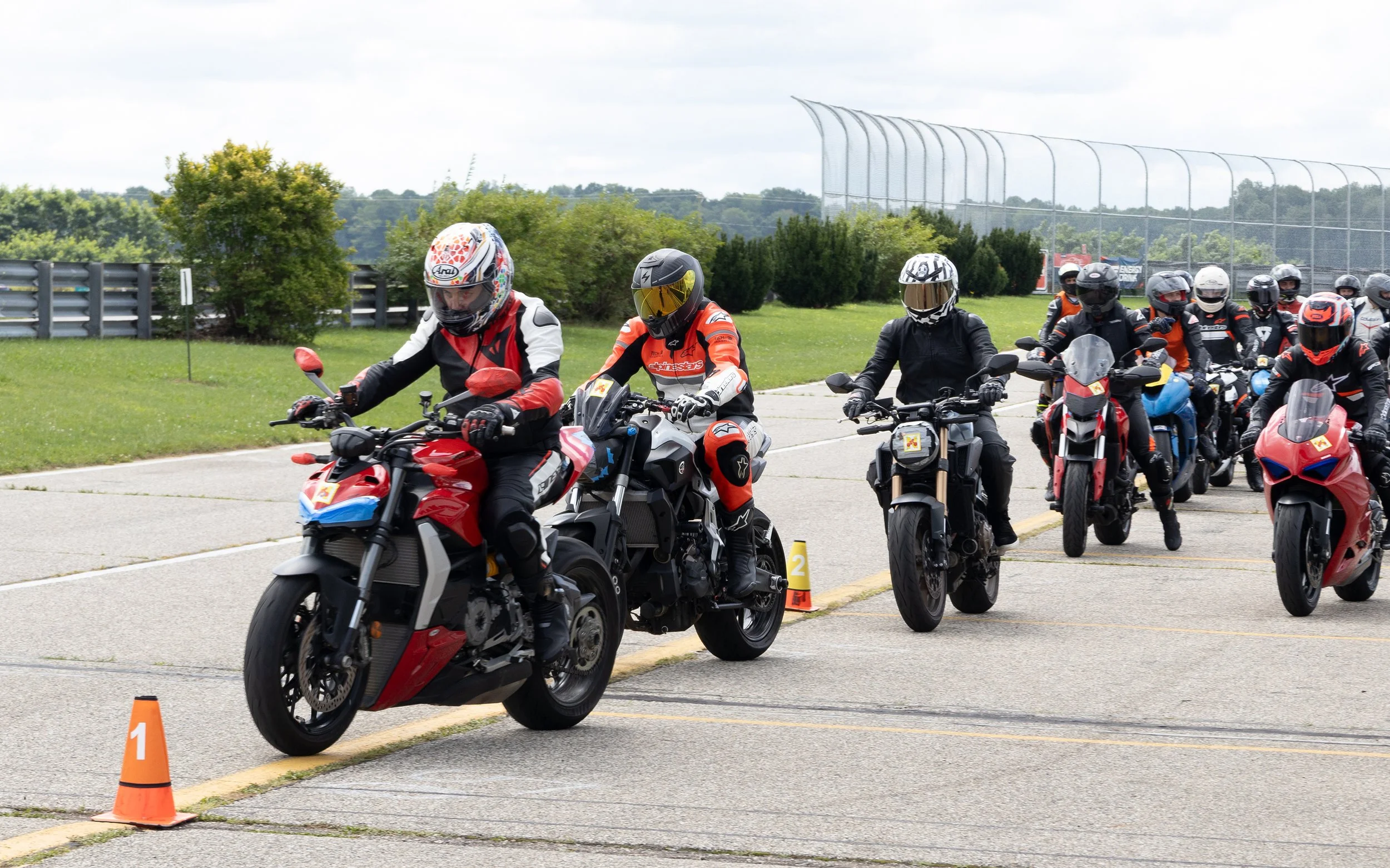 Group of motorcyclists in full gear riding on a track, some with helmets with reflective visors, during a motorcycle training or racing event.