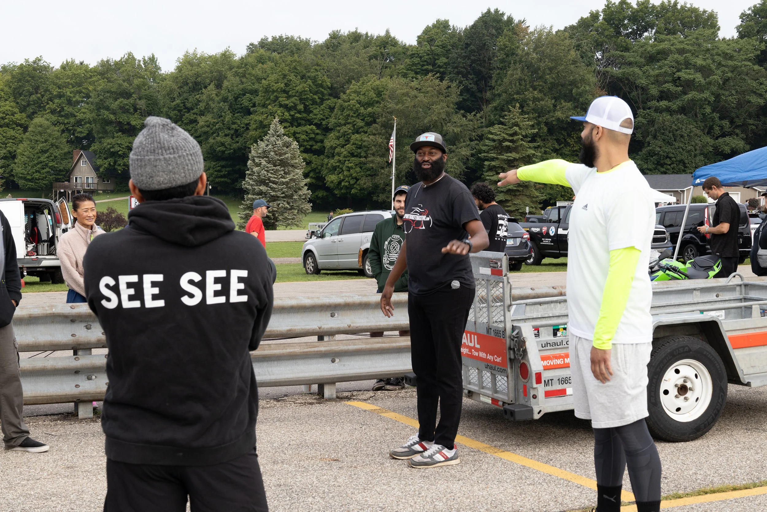 People engaging in conversations outdoors near vehicles, with trees and houses in the background.