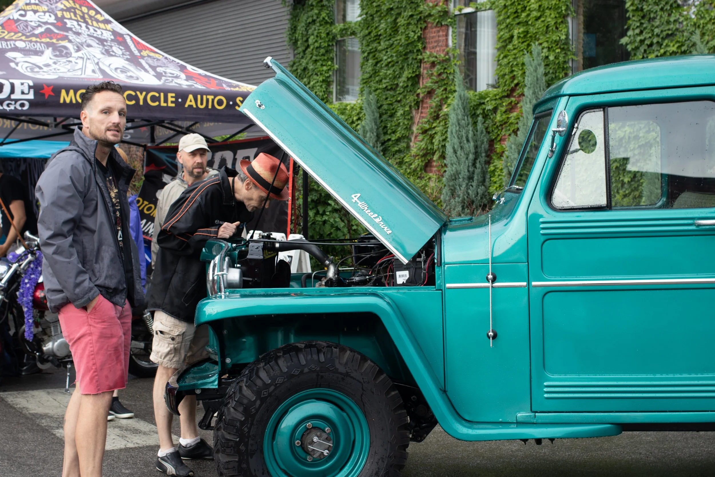 People inspecting a teal vintage truck with the hood open at an outdoor event, with an event tent and green foliage in the background.