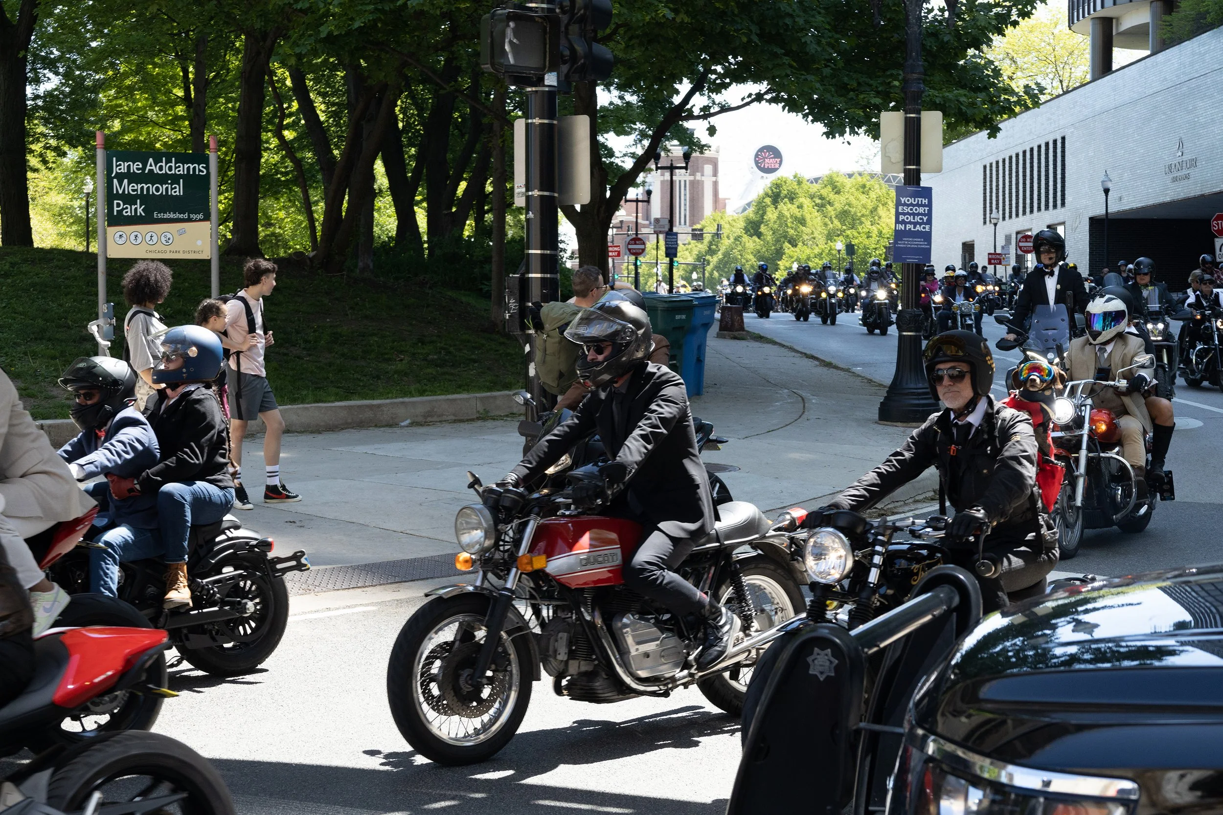 Bikers and pedestrians at the entrance to Jane Addams Memorial Park, with a crowd of motorcyclists riding through, trees and buildings in the background, and signs displaying park and policy information.