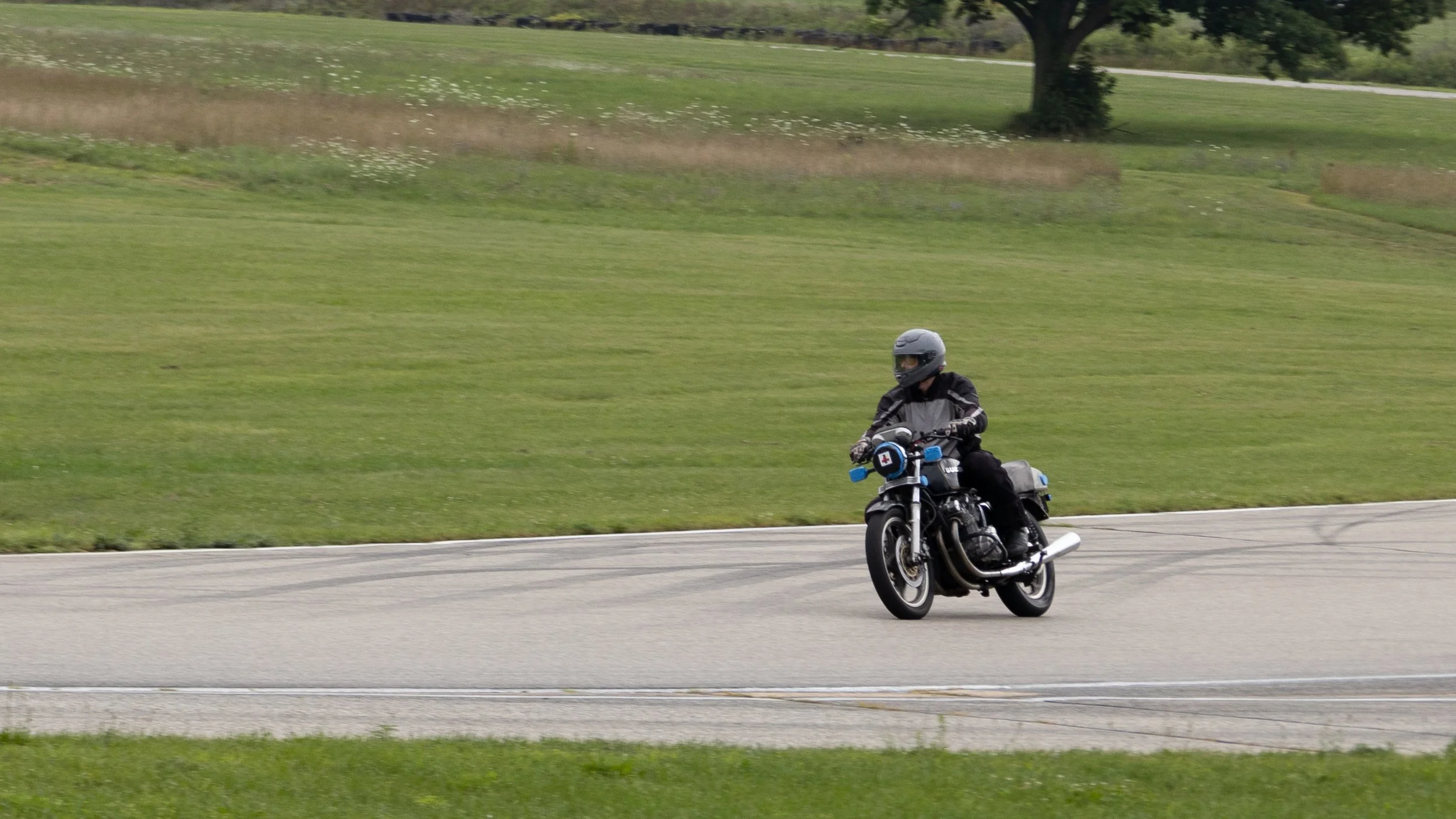A person riding a motorcycle on a paved road with grassy fields and trees in the background.