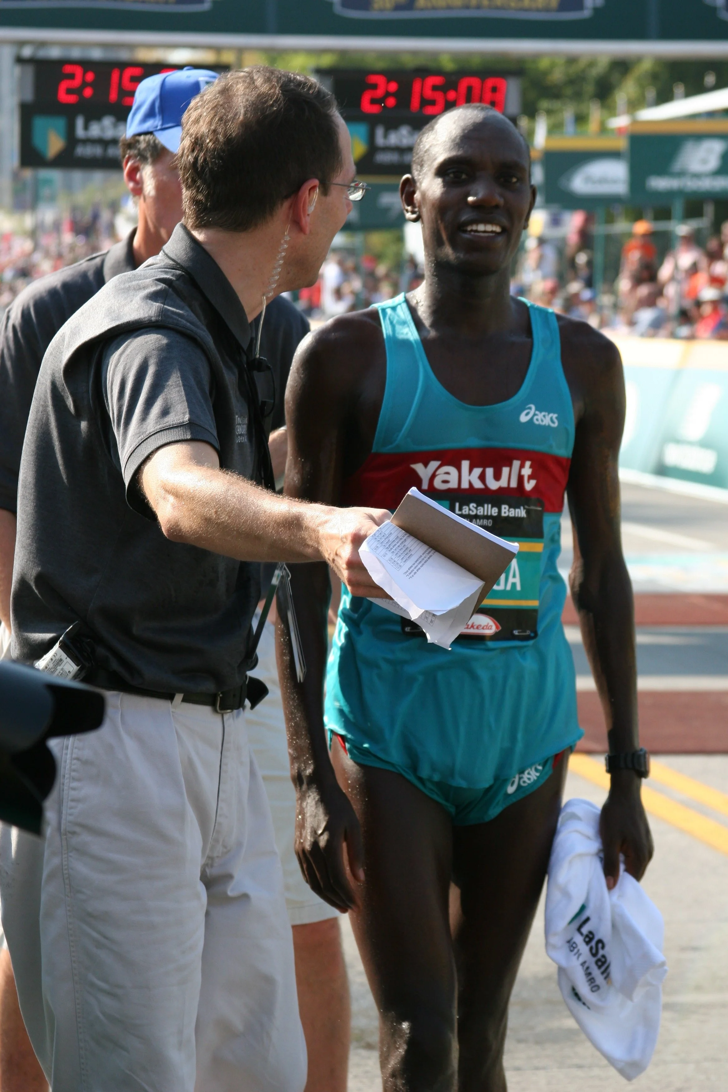 A male marathon runner with dark skin, wearing a turquoise and red Asics tank top with Yakult sponsorship, is being interviewed or assisted by officials at the finish line of a race, with a crowd in the background and digital timers overhead.