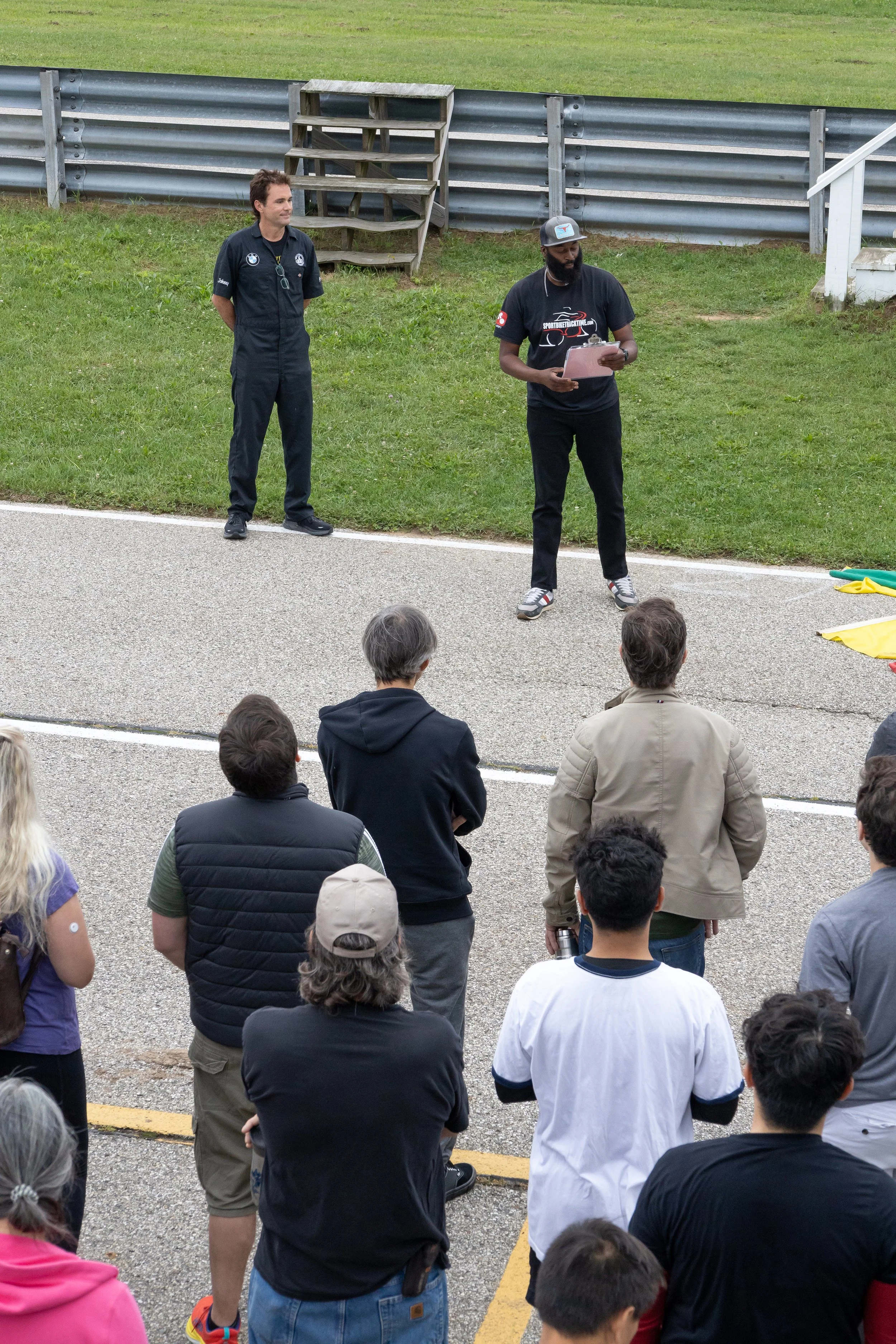Two men standing in front of a group of seated people at an outdoor event on a racetrack, with a grassy area and metal barriers in the background. One is wearing a black shirt with logos, holding a clipboard, and the other is in a black jumpsuit with