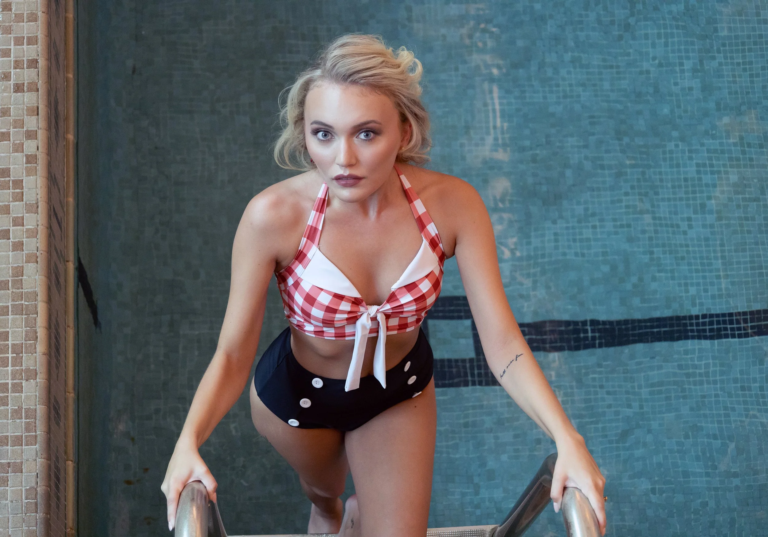 A woman with blonde hair and blue eyes, wearing a red and white checkered swimsuit top and black shorts, sitting on the stairs of a swimming pool, holding the metal handrails, looking at the camera.