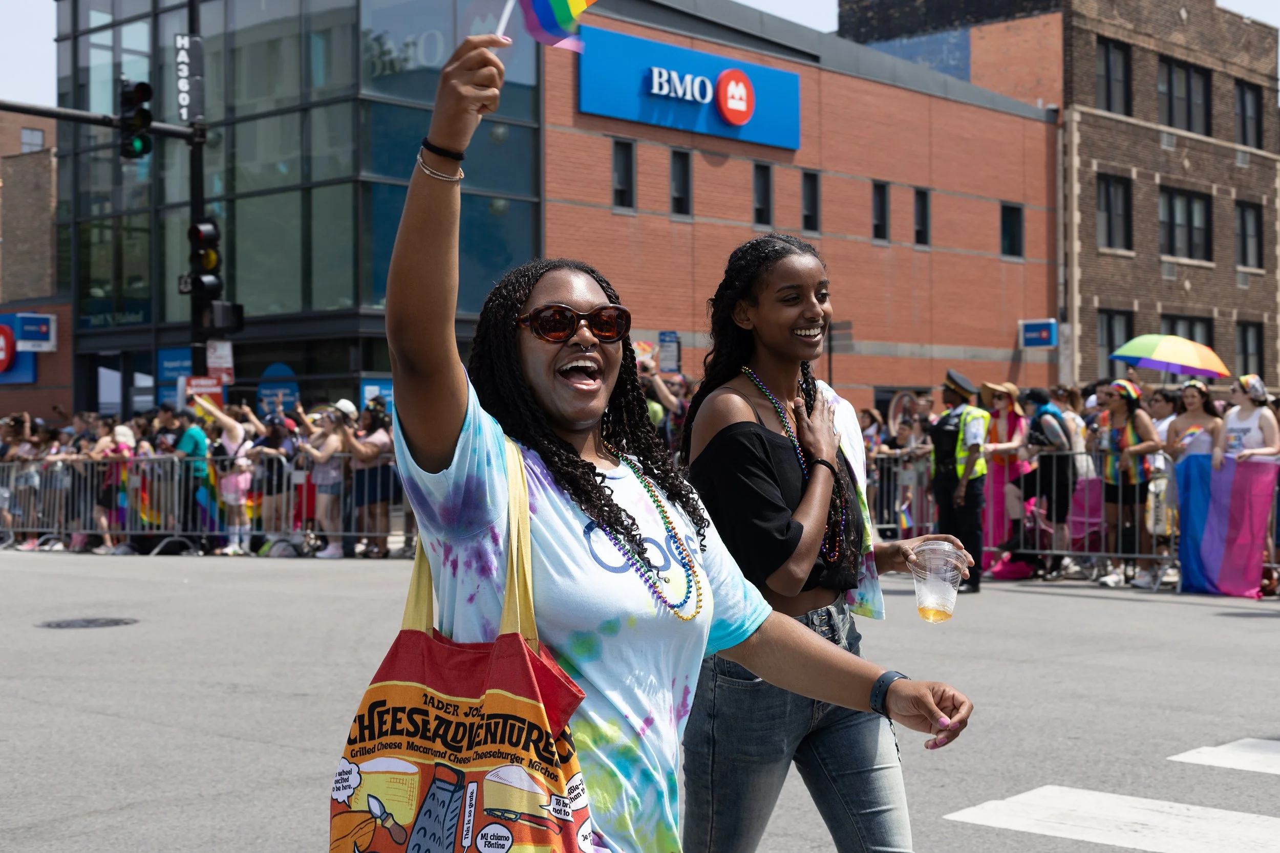 Two women participating in a parade, smiling and celebrating. One woman is wearing colorful tie-dye clothing and sunglasses, waving a small rainbow flag, while the other woman is holding a drink, both surrounded by a crowd of spectators and parade pa