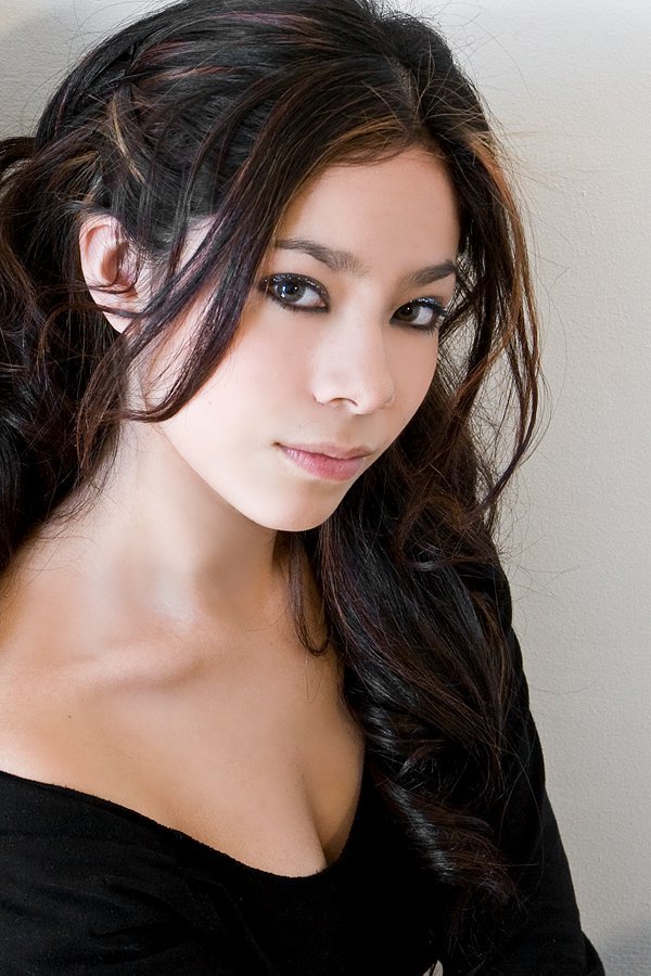 A woman with long, wavy dark hair and fair skin looking at the camera with a neutral expression, wearing a black top against a plain light-colored wall.