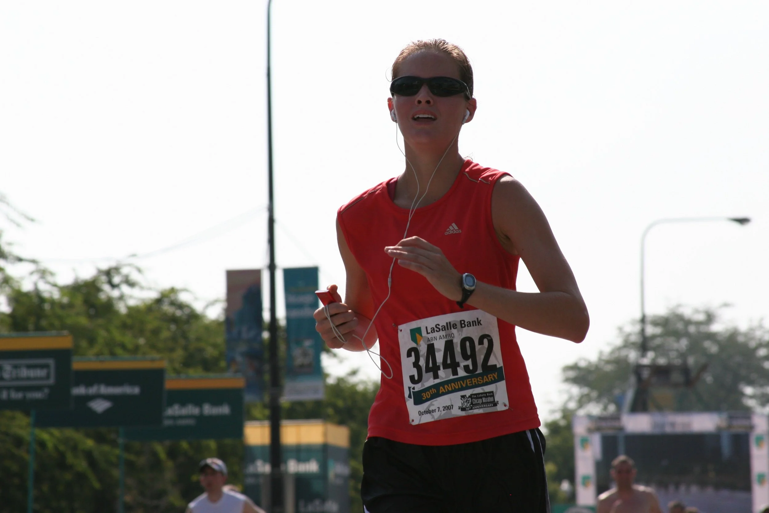 A woman running in a marathon, wearing sunglasses, a red athletic tank top, and a bib with the number 34492, holding a smartphone and listening to music with earphones.