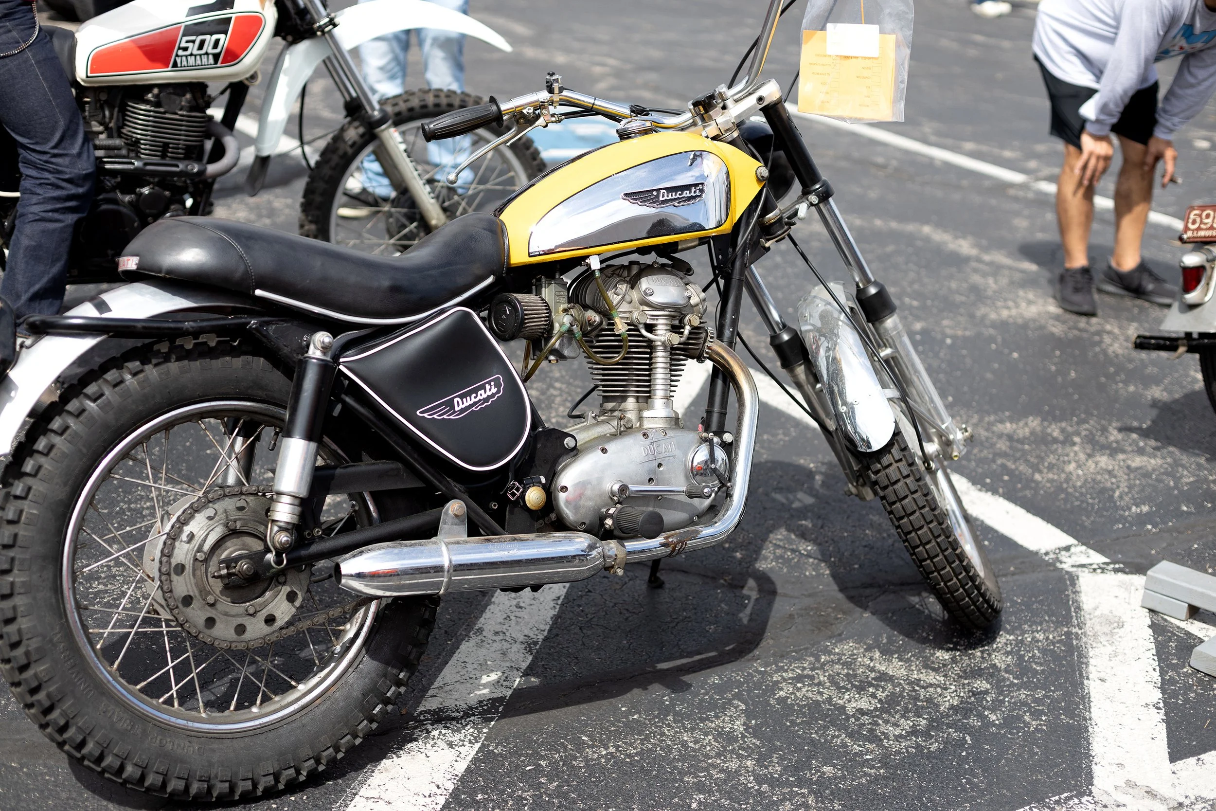A vintage yellow and black Ducati motorcycle parked in a parking lot during a motorcycle show, with people inspecting other bikes in the background.