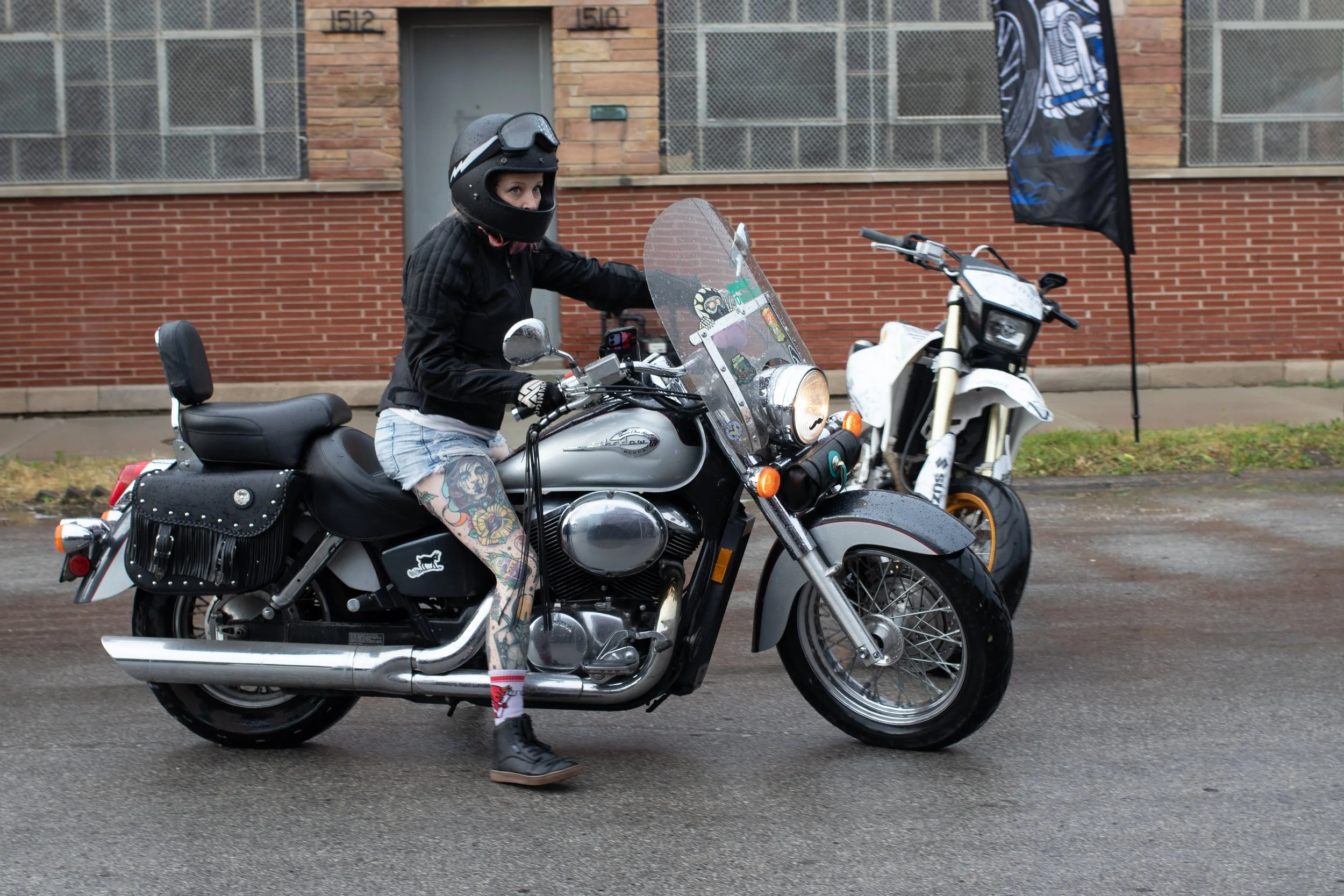 A woman riding a motorcycle outdoors, wearing a black helmet, black jacket, and shorts, with colorful tattoos on her legs, parked on the street near a brick building.