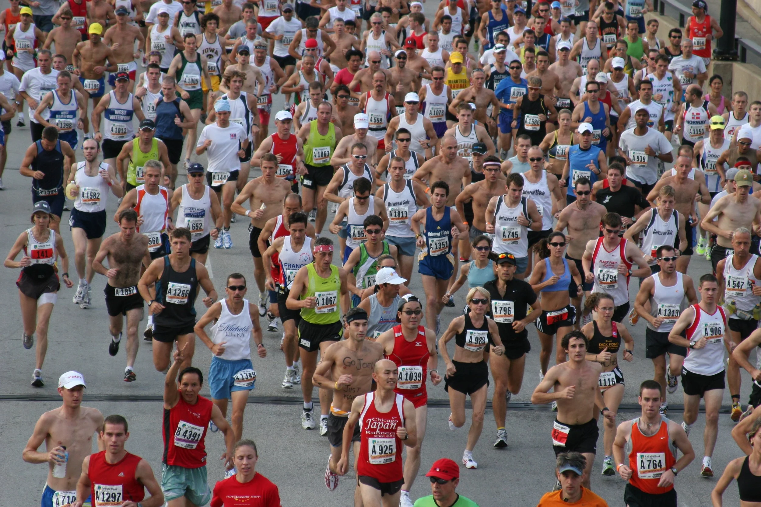 Large group of marathon runners participating in a race, some shirtless, with various bib numbers and athletic clothing, running on a city street.