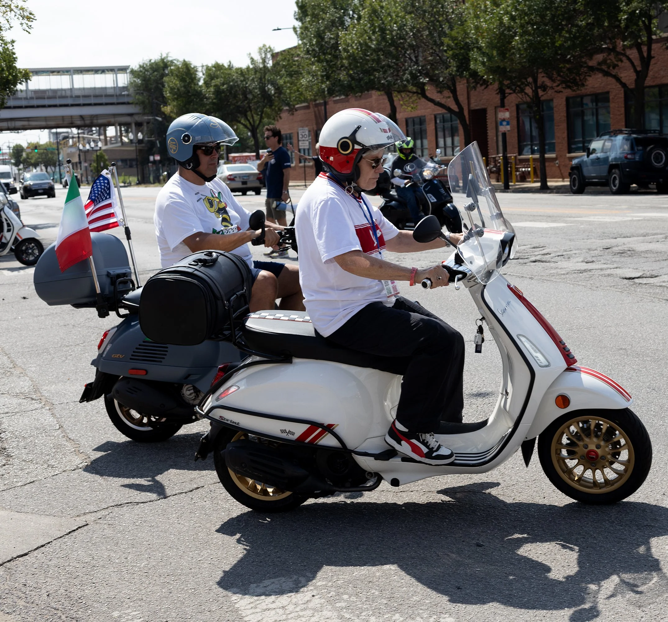 Two people wearing helmets riding a scooter with flags attached, crossing a city street on a sunny day.