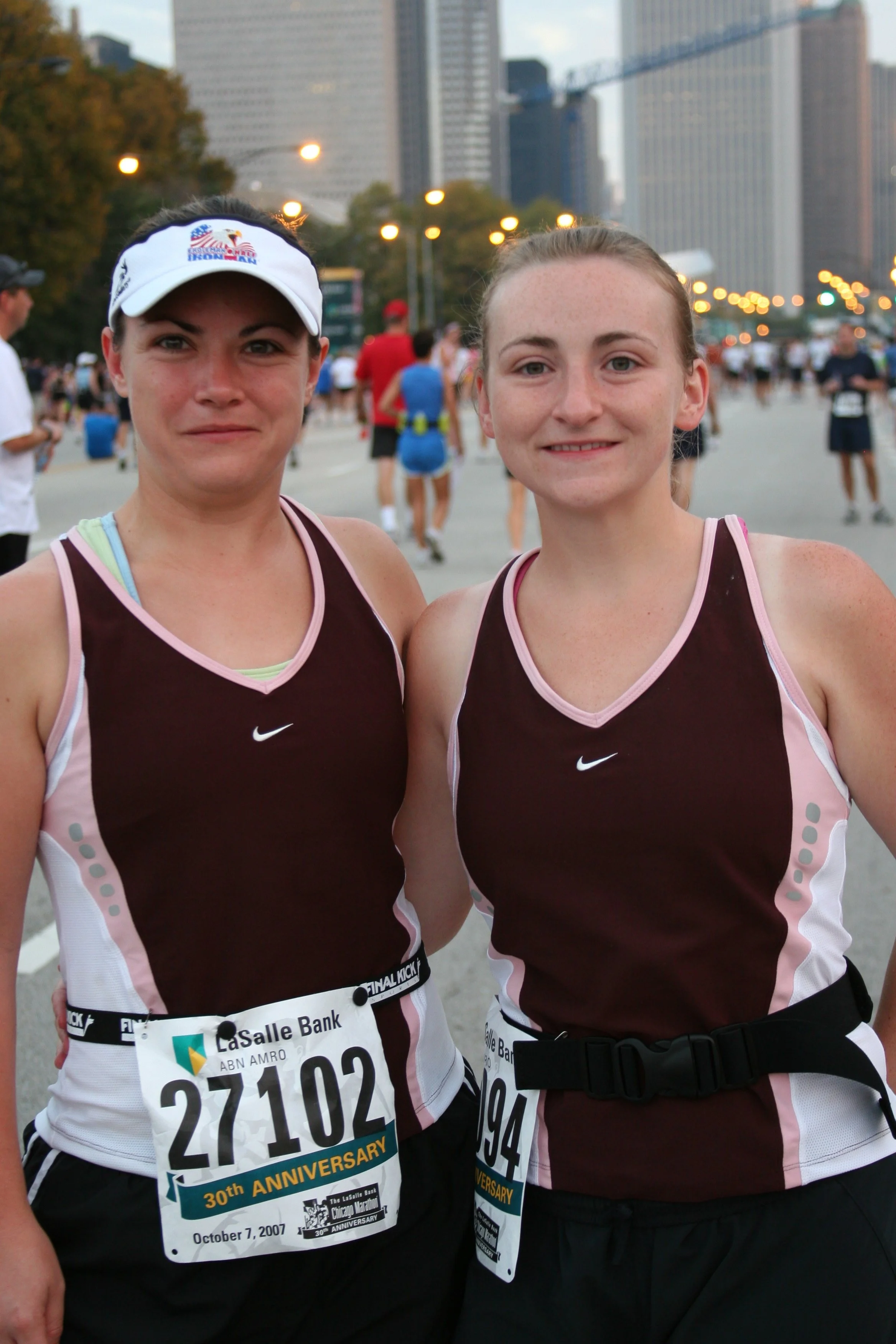 Two women in running attire with race bibs, standing outdoors in a city with tall buildings in the background, after participating in a marathon, smiling at the camera.