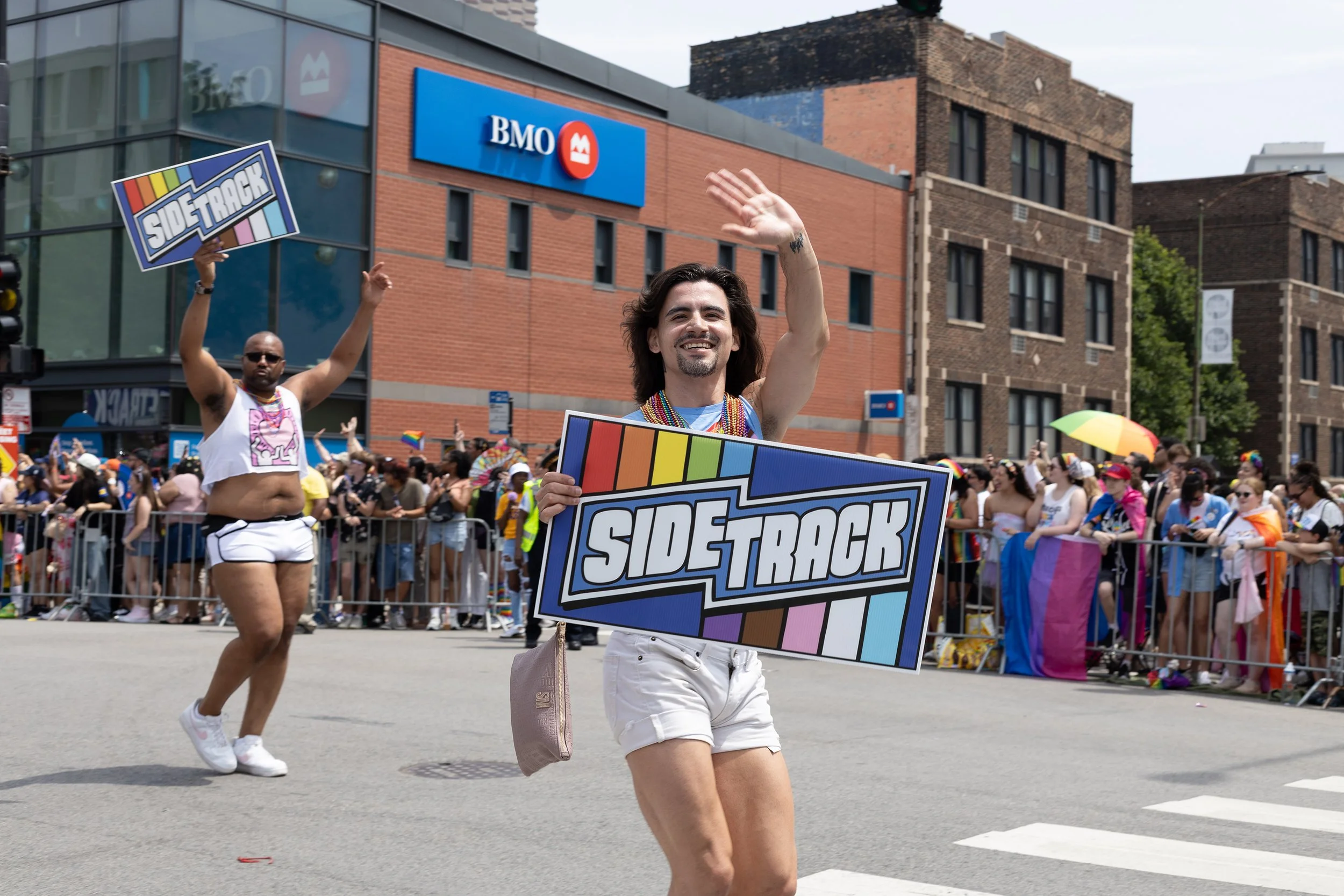 Two individuals proudly participate in a pride parade. The person in front is smiling, wearing white shorts, a beaded necklace, and a shirtless top with a rainbow-themed sign reading 'SIDE TRACK'. The person behind is also holding a similar sign and 