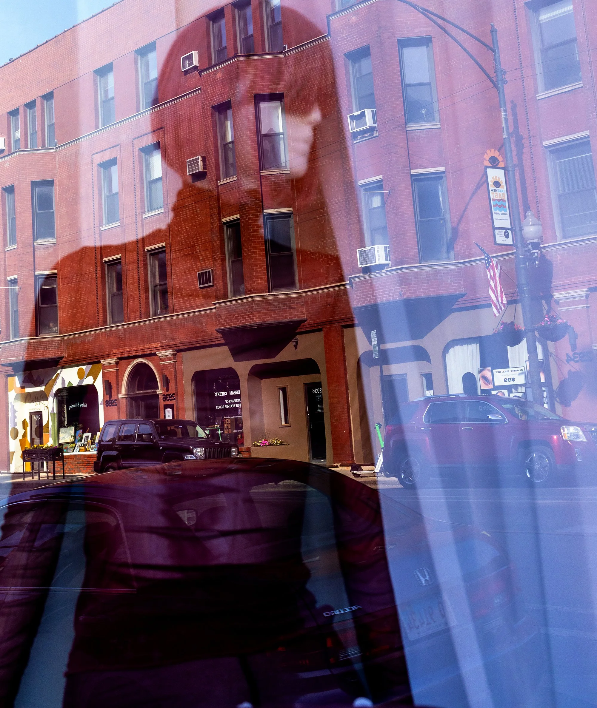 Reflections of red brick buildings, storefronts, a black car, and a street scene in the glass window of a storefront on a sunny day.