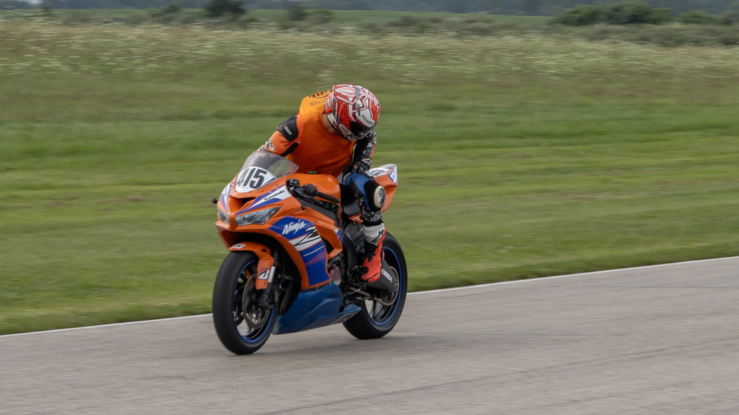 Motorcycle rider leaning into a turn on a race track, wearing orange and black racing gear and a red and white helmet, riding an orange and blue Kawasaki motorcycle.
