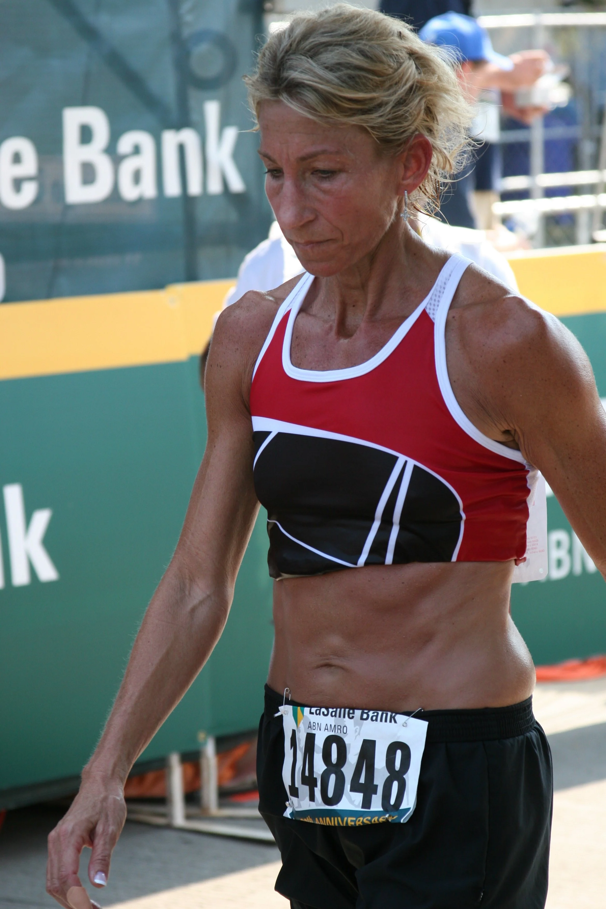 A female marathon runner in a red, white, and black athletic outfit with a bib number 14848, walking after a race.