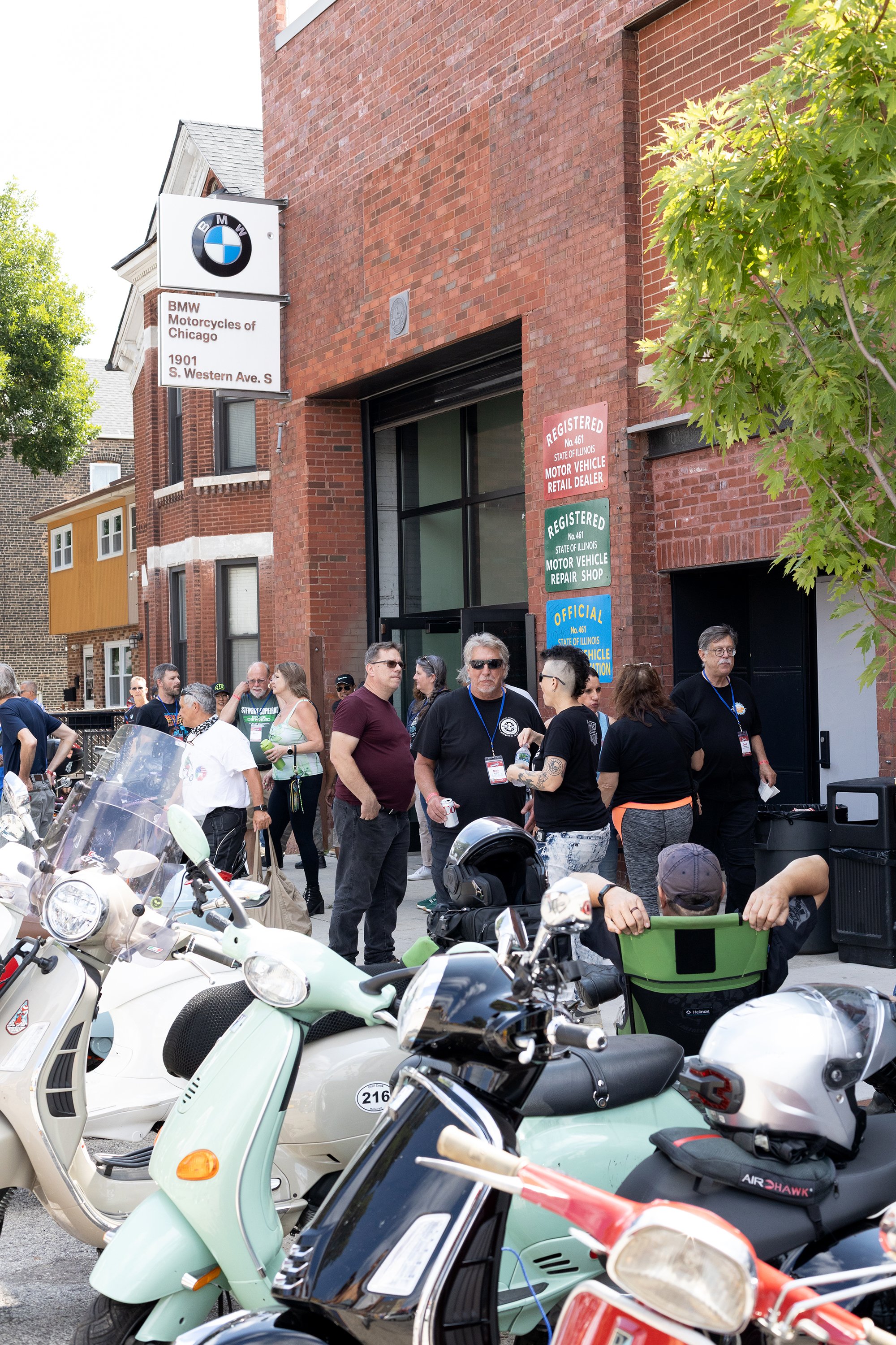People gathered outdoors near a brick building with BMW Motorcycles of Chicago sign. Several motorcycles and scooters parked in foreground. Green leafy tree on right side.