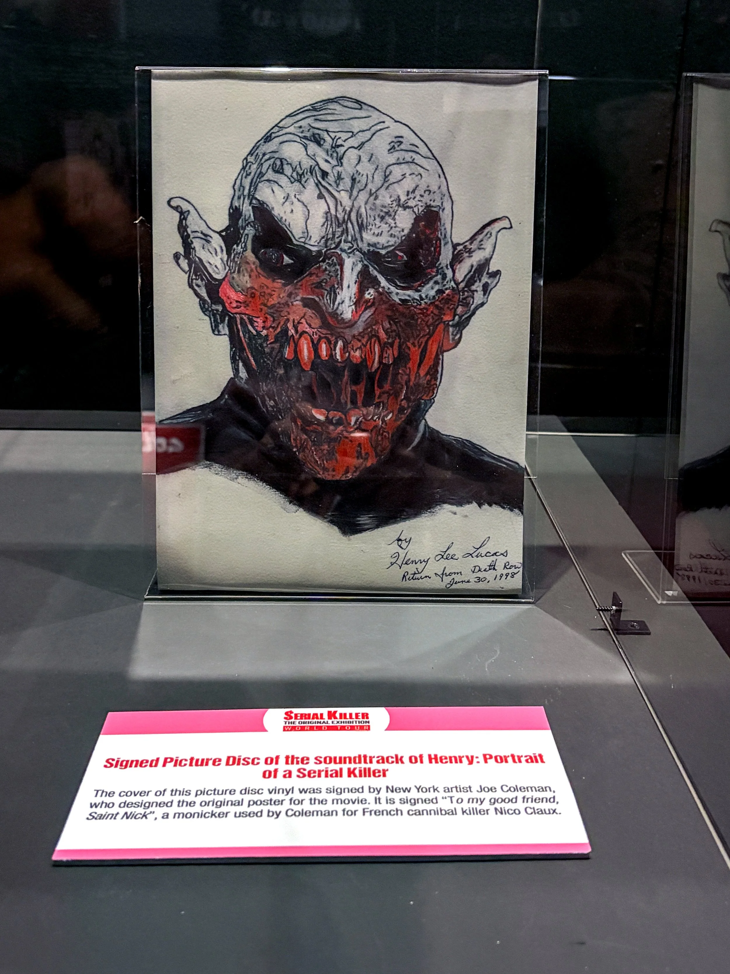 A vinyl record cover with a black and white sketch of a grotesque, zombie-like face with exaggerated features and red accents, displayed in a case at an exhibit. A pink informational placard below describes it as a signed picture disc of the soundtra
