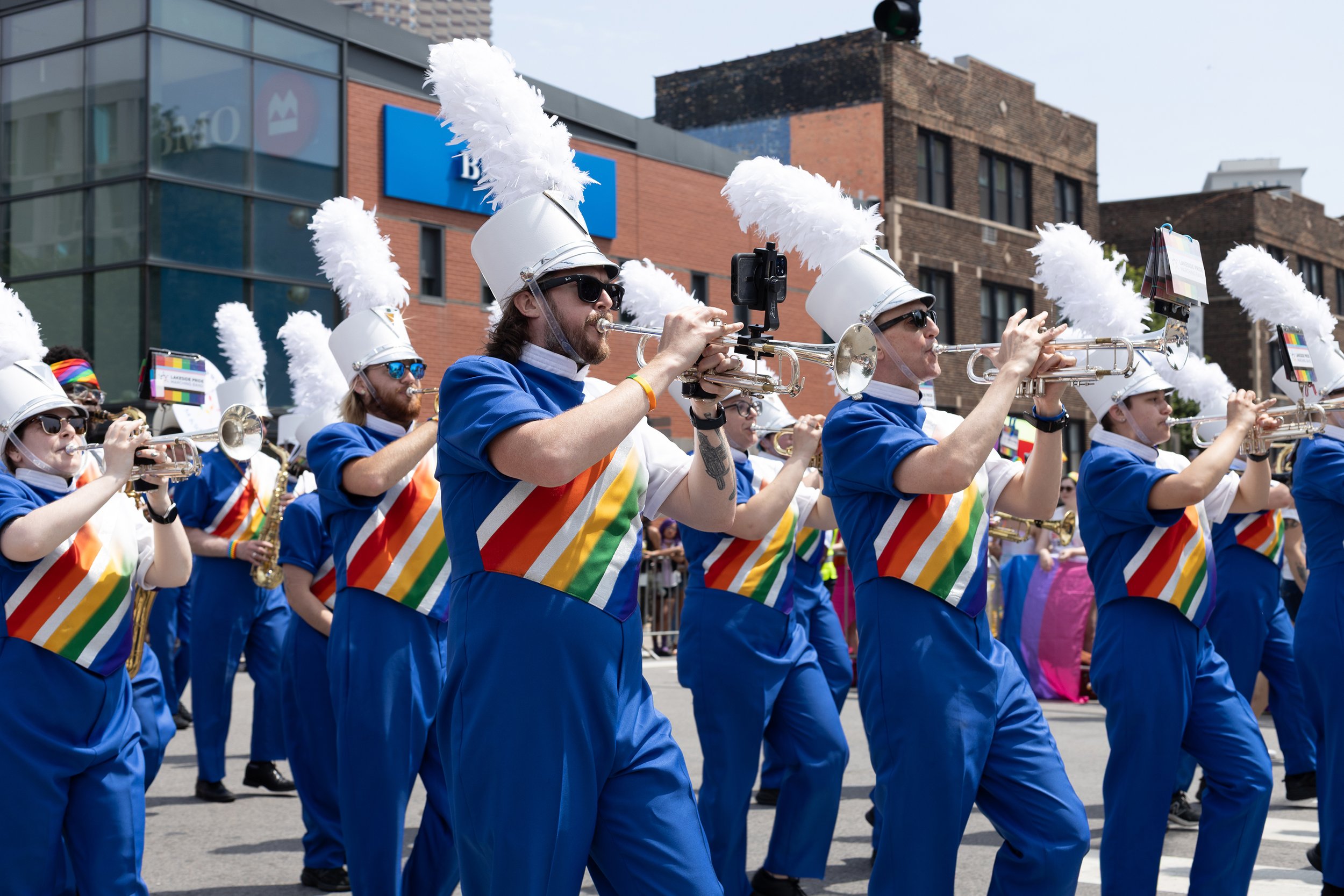 A marching band parade featuring members wearing blue uniforms with rainbow-striped sashes and white hats with large white feathers, playing trumpets as they march down the street.