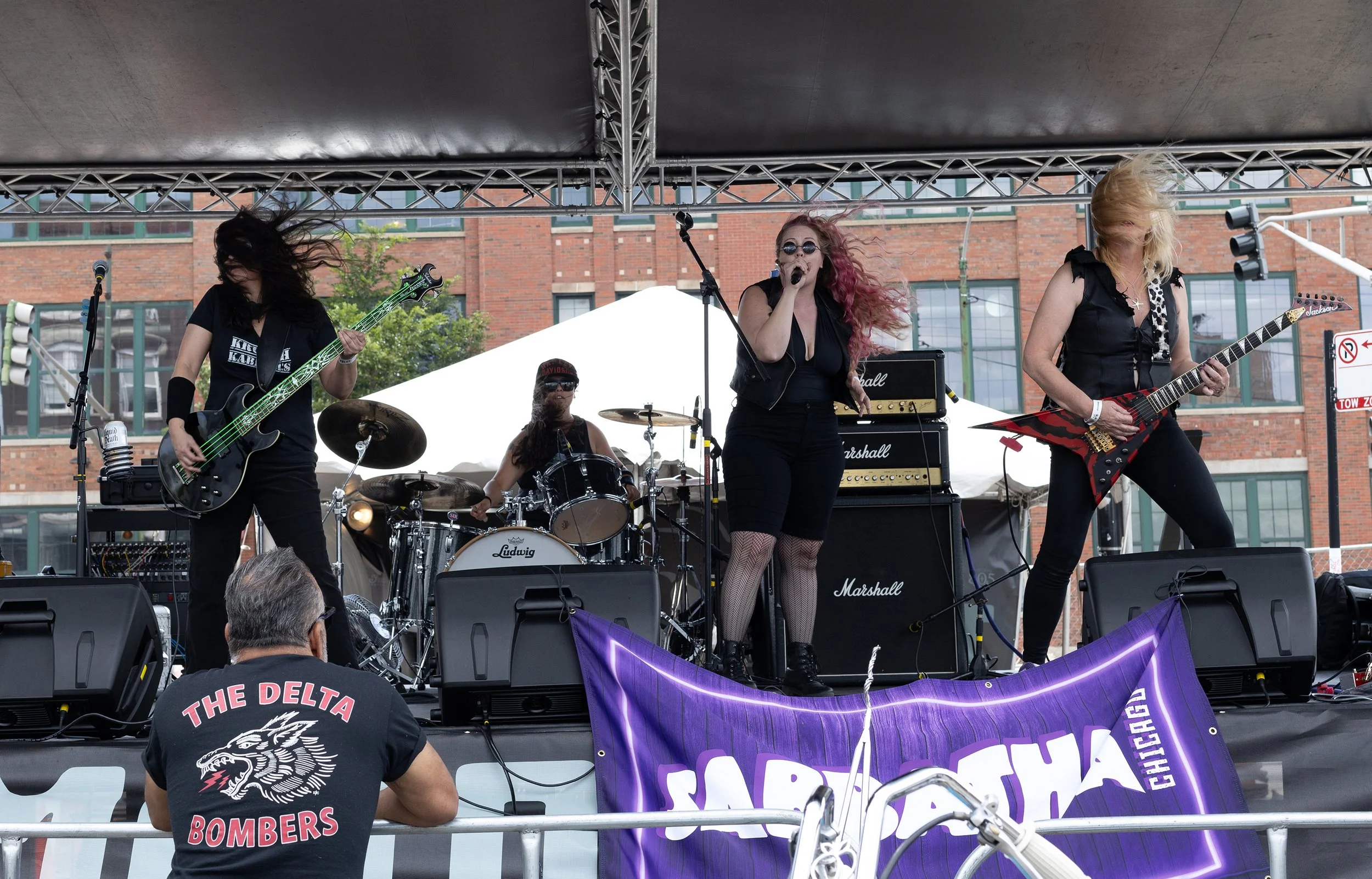 A live outdoor band performance with female singers playing electric guitar and drums, with a brick building in the background and people in the foreground.