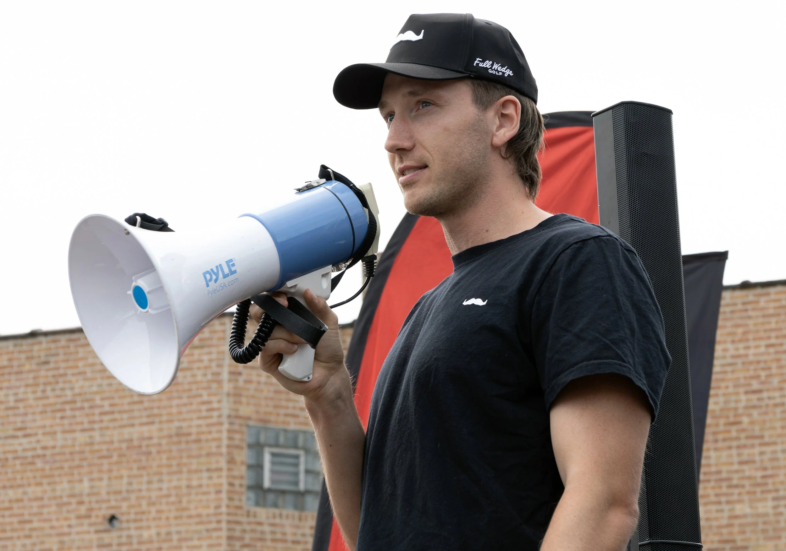 A young man wearing a black cap and matching T-shirt holds a blue and white megaphone with the word 'DYLE' on it, standing outdoors in front of a brick building with windows.