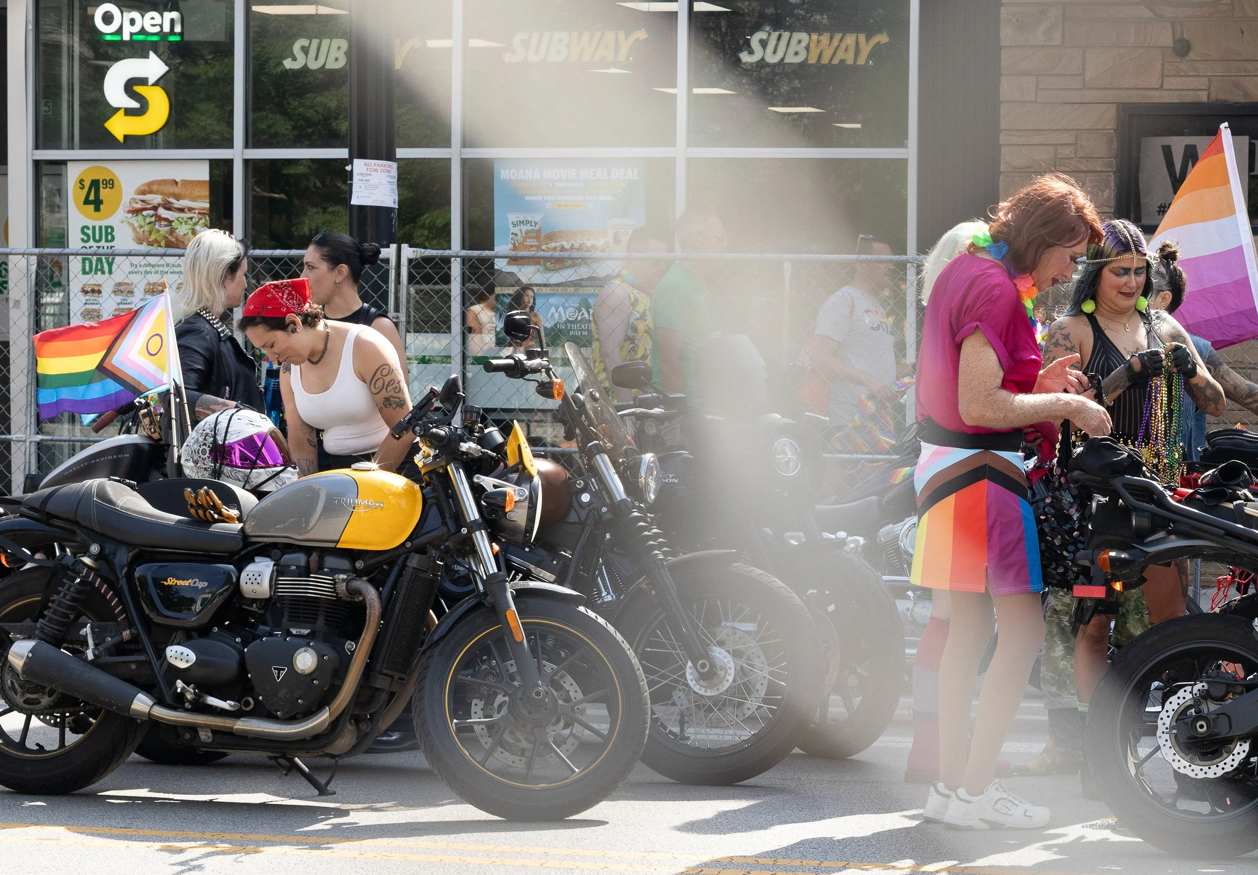 A group of people dressed in colorful, rainbow-themed clothing and accessories gathered outside a Subway restaurant, with motorcycles parked in the foreground.