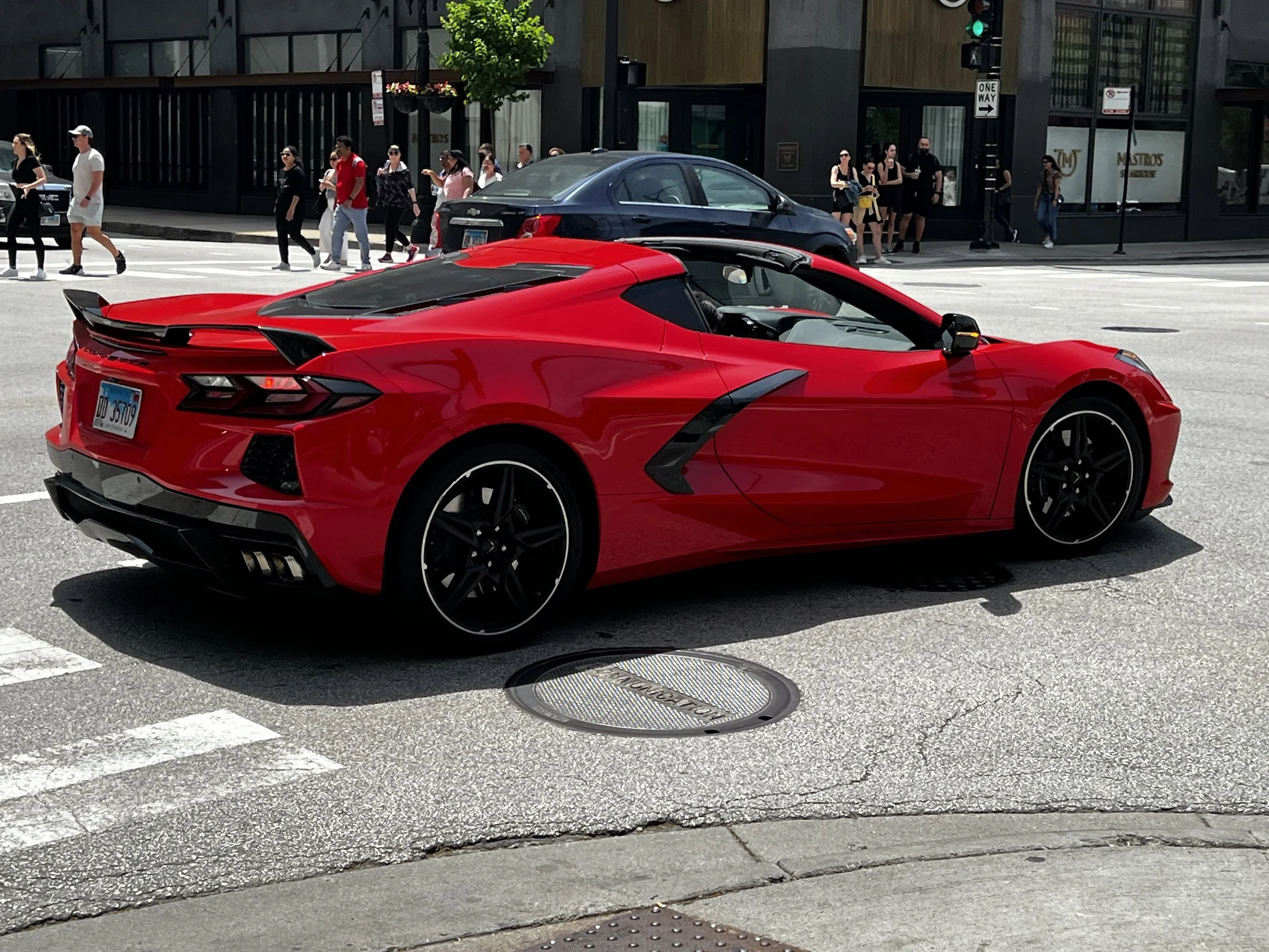 A red sports car is driving past a group of pedestrians crossing the street in an urban setting. The pedestrians are waiting on the sidewalk, and the background features a building with large windows and signs, including a green traffic light visible